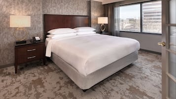 Quarto de hotel com uma cama grande, duas mesas de cabeceira com abajures, uma cabeceira de madeira e vista para edifícios através de uma janela.