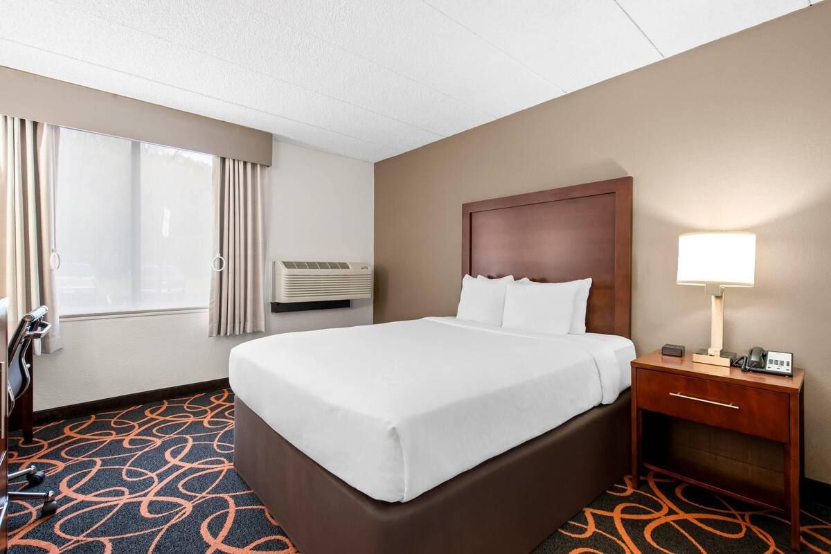 A hotel room with a large bed, a bedside table with a lamp, an air conditioning unit, and a window with curtains.