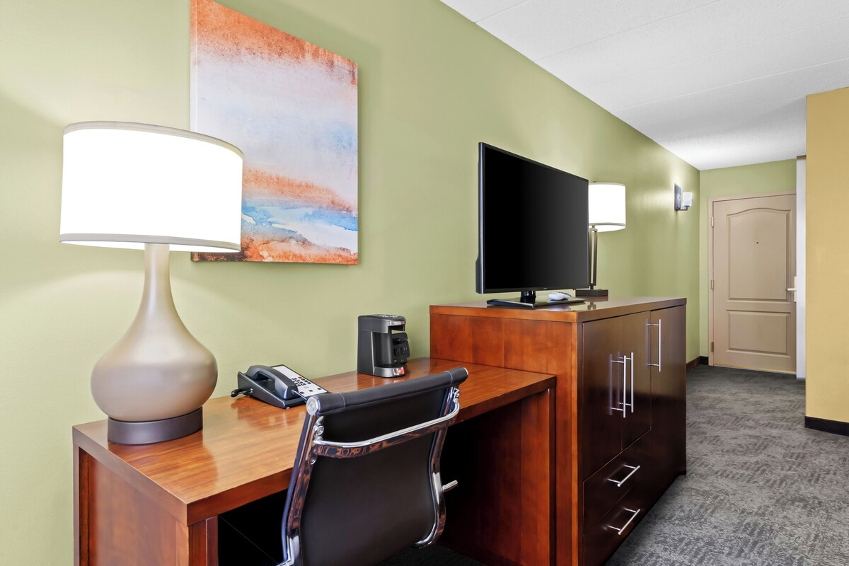 A hotel room with a desk, chair, lamp, coffee maker, telephone, television, and artwork on the wall.
