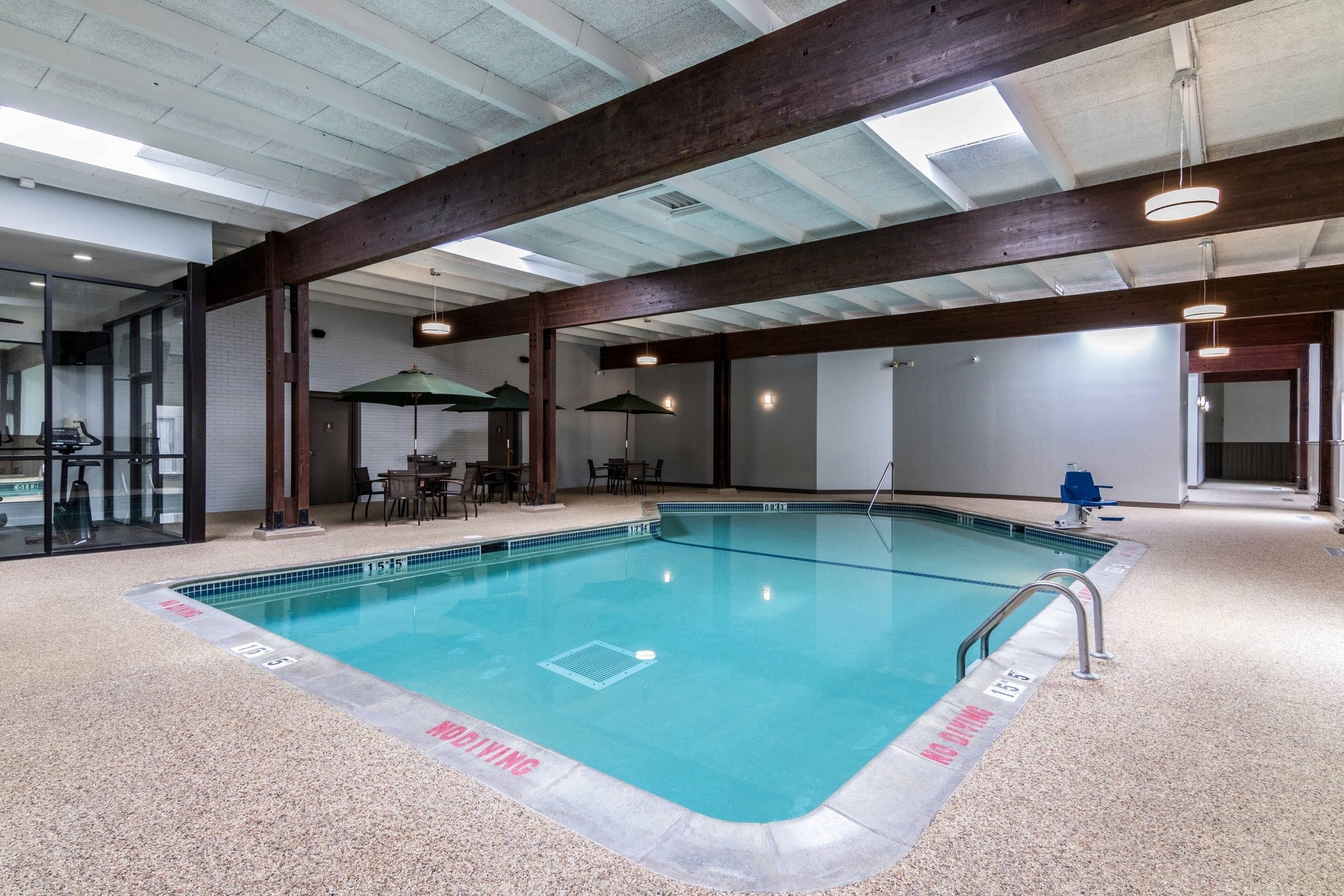 An indoor swimming pool with clear water, surrounded by a tiled floor, wooden beams, and a glass enclosure.