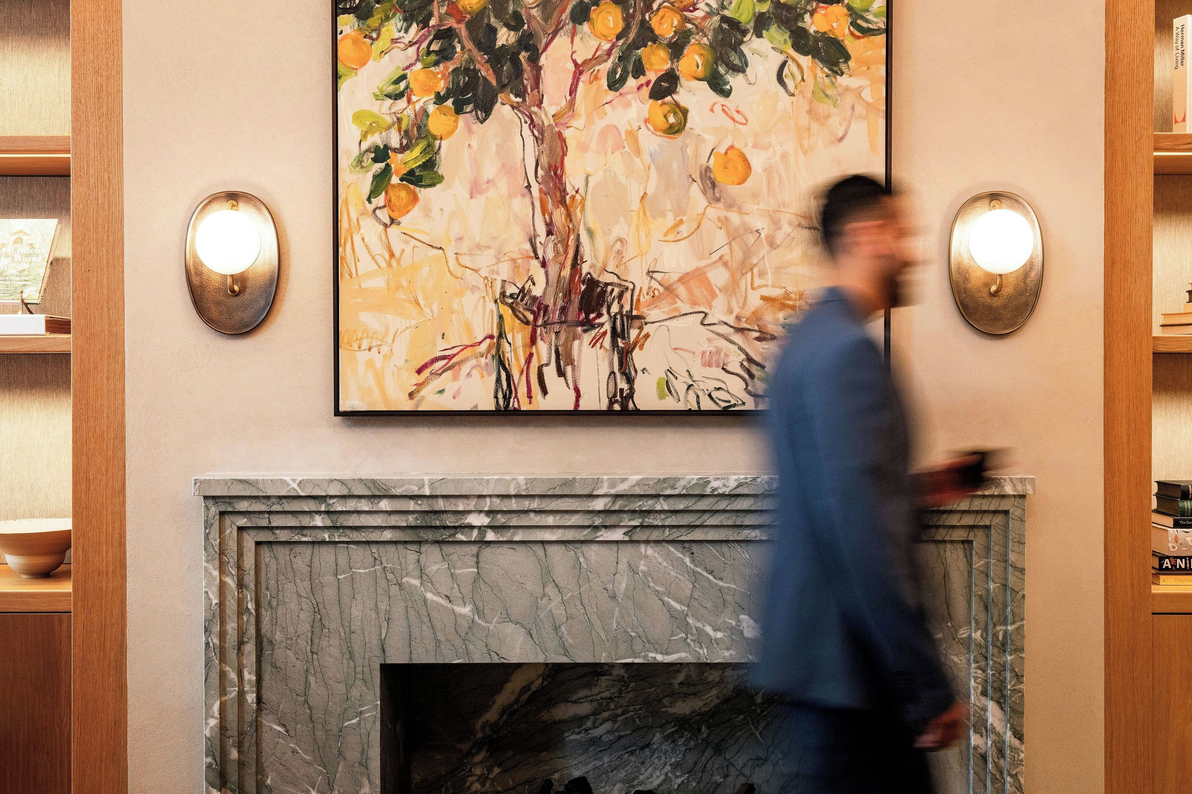 A room with a marble fireplace, a painting of a tree with oranges, and two wall sconces.