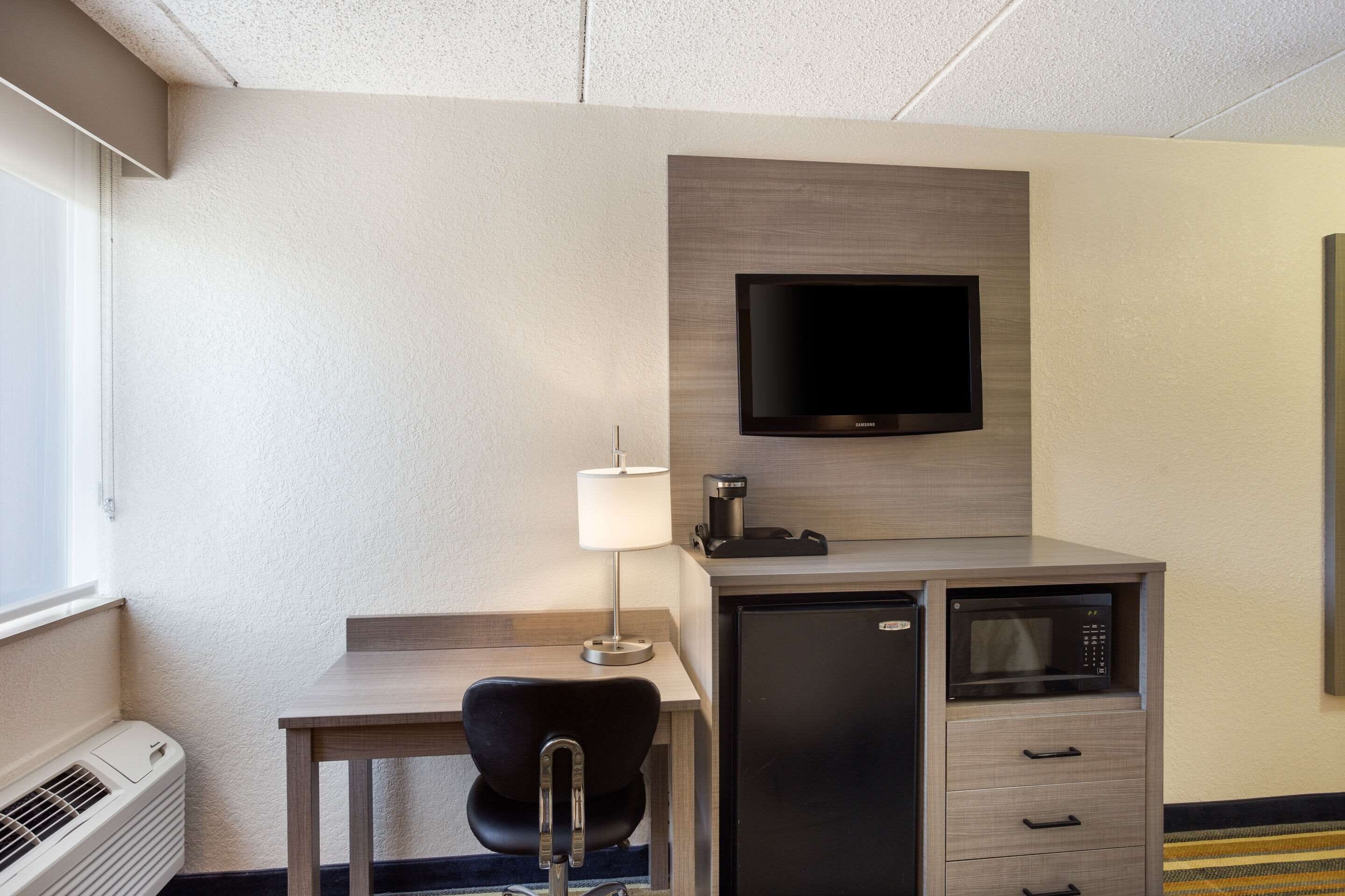 Een compacte hotelkamer met een bureau, stoel, flatscreen televisie, minibar, magnetron en een lamp.