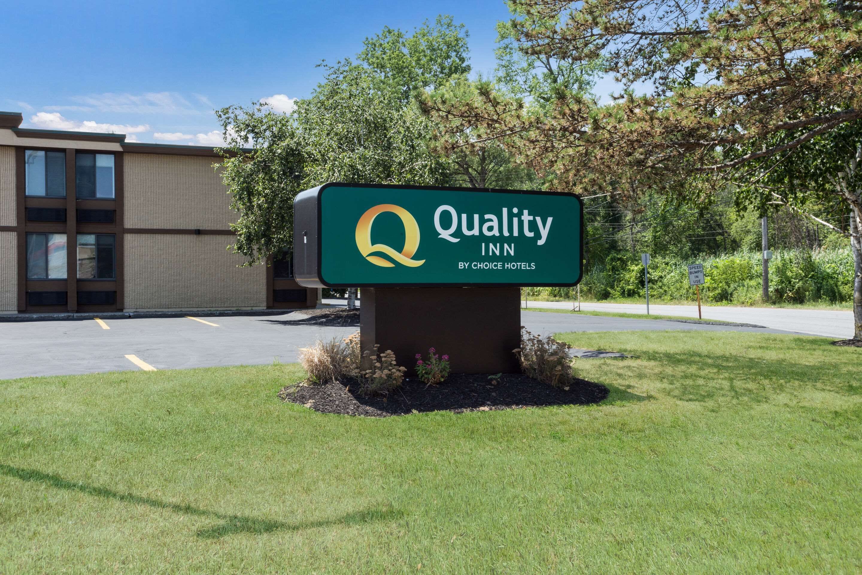 A Quality Inn sign with a logo, set in a landscaped area with grass and shrubs, in front of a building with a modern design.