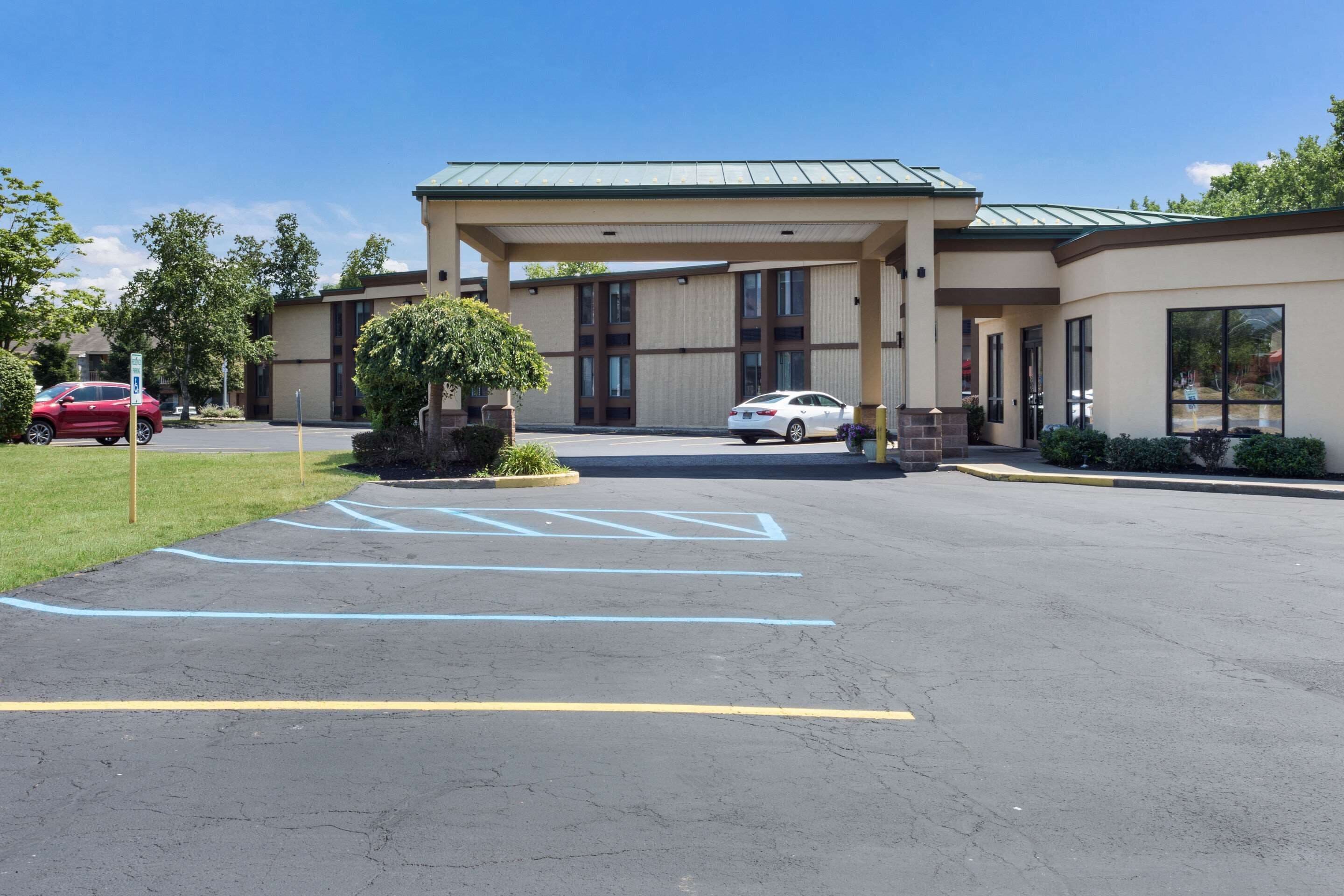 A hotel entrance with a covered parking area, marked parking spaces, and a small landscaped area with a bush.