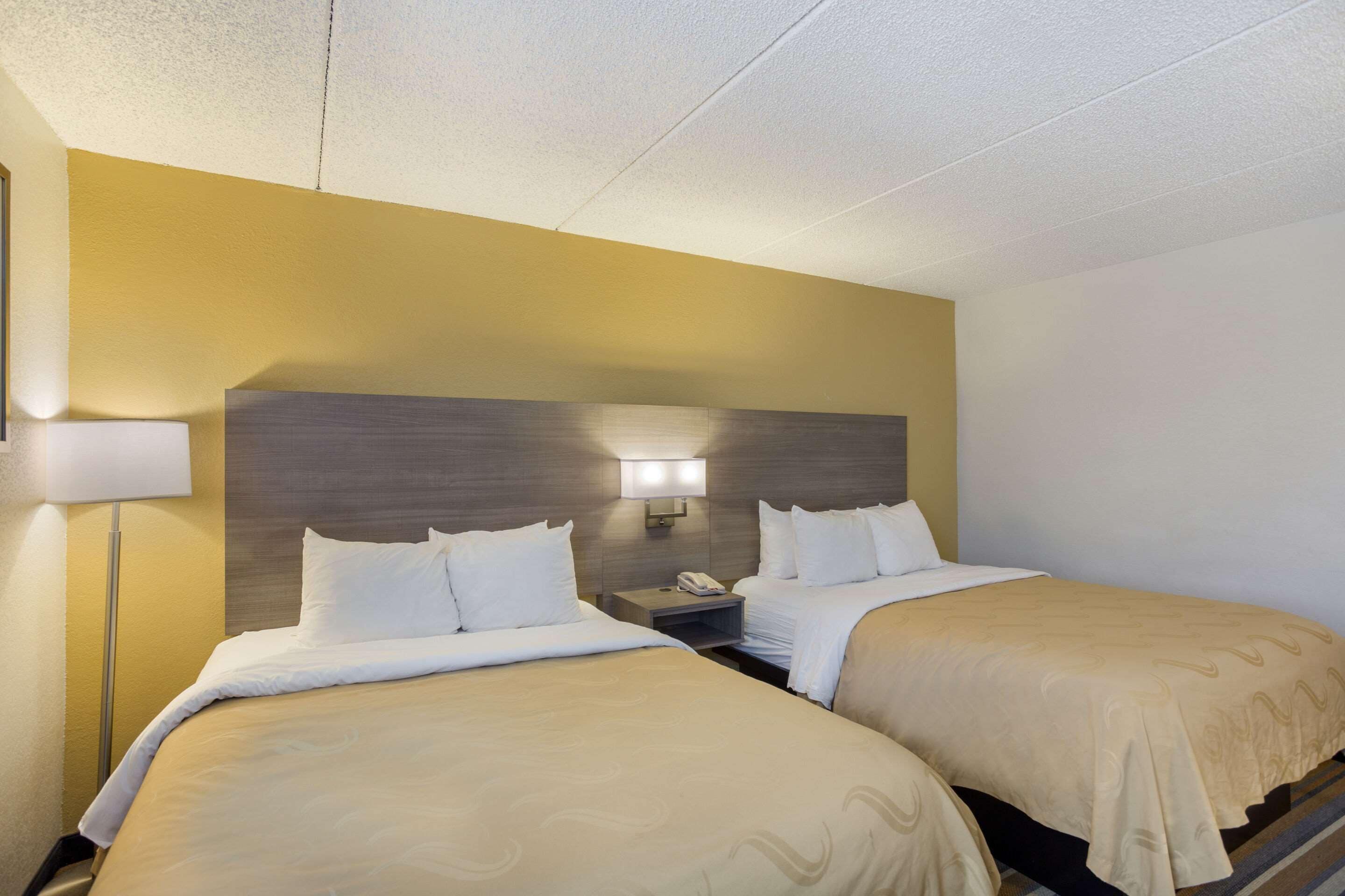 A hotel room with two beds, a nightstand with a lamp, and a wall-mounted light fixture.
