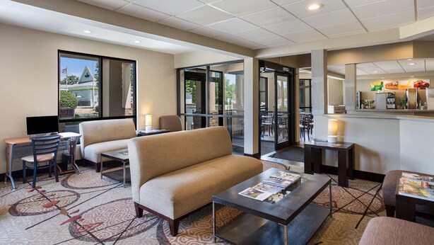 A hotel lobby with a sofa, coffee table, and seating area. There is a desk with a computer and a chair. The lobby has a patterned carpet and a glass door leading outside. - Quality Inn Albany Colonie (Albany)