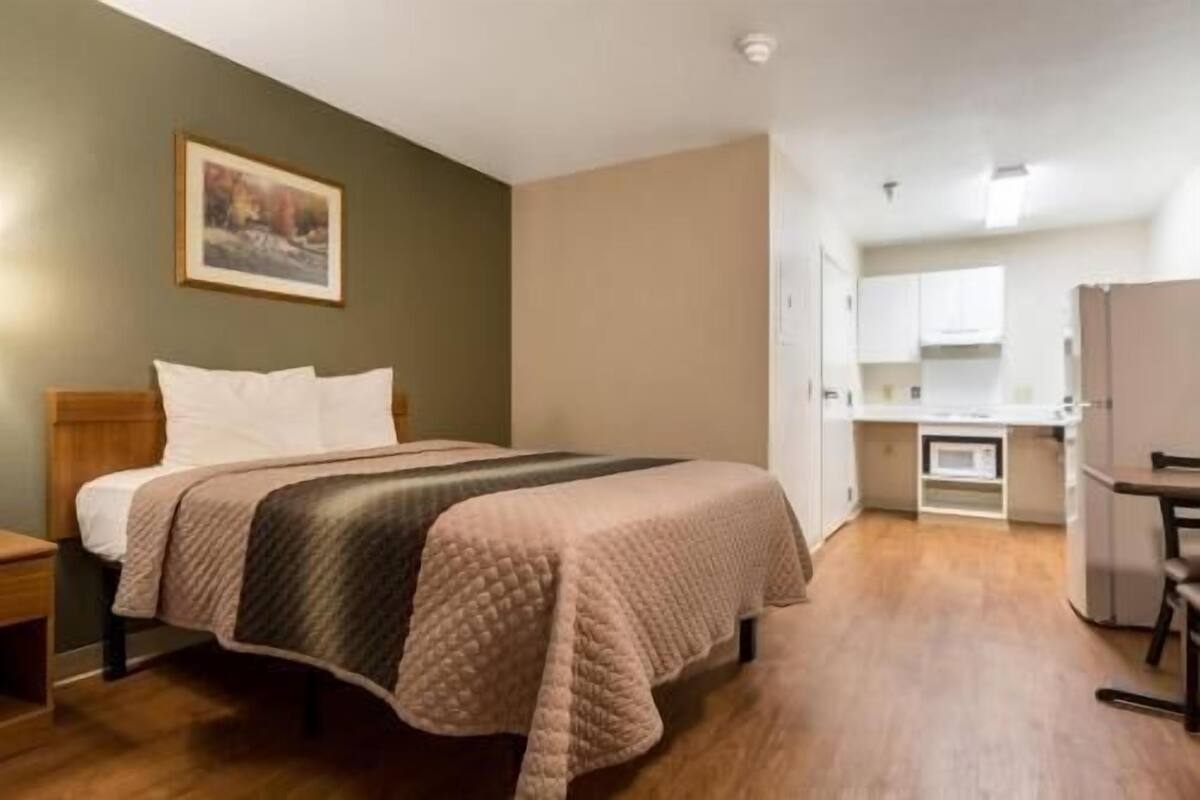 Ein Hotelzimmer mit einem Bett, einem Nachttisch, einem Bild an der Wand und einer Küchenzeile im Hintergrund.