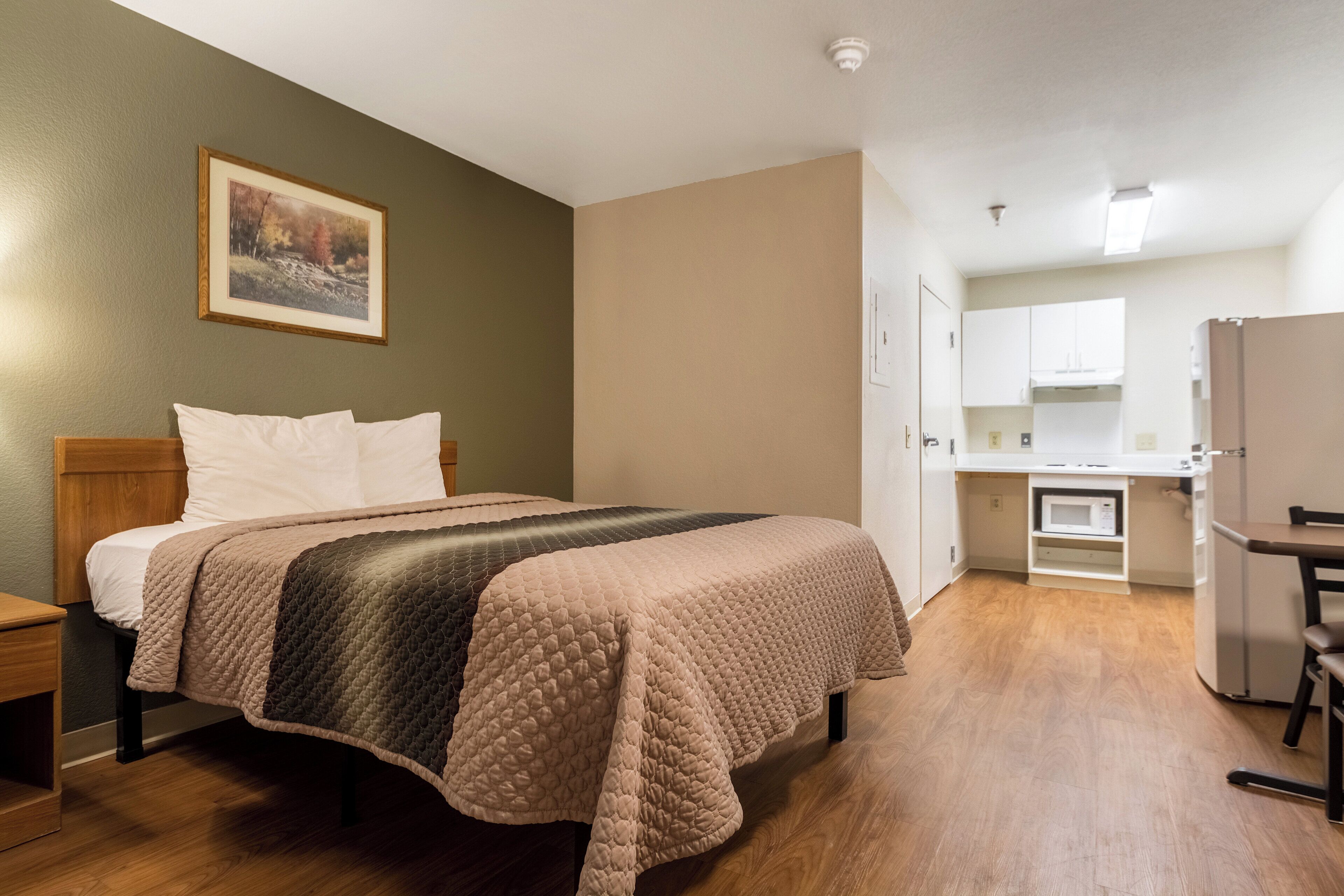 Una habitación de hotel con una cama, una mesita de noche, un cuadro en la pared y una kitchenette al fondo.