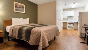 Una habitación de hotel con una cama, una mesita de noche, un cuadro en la pared y una kitchenette al fondo.