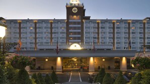 A large hotel with a prominent clock tower, a well-lit entrance, and a landscaped driveway leading to the parking area.