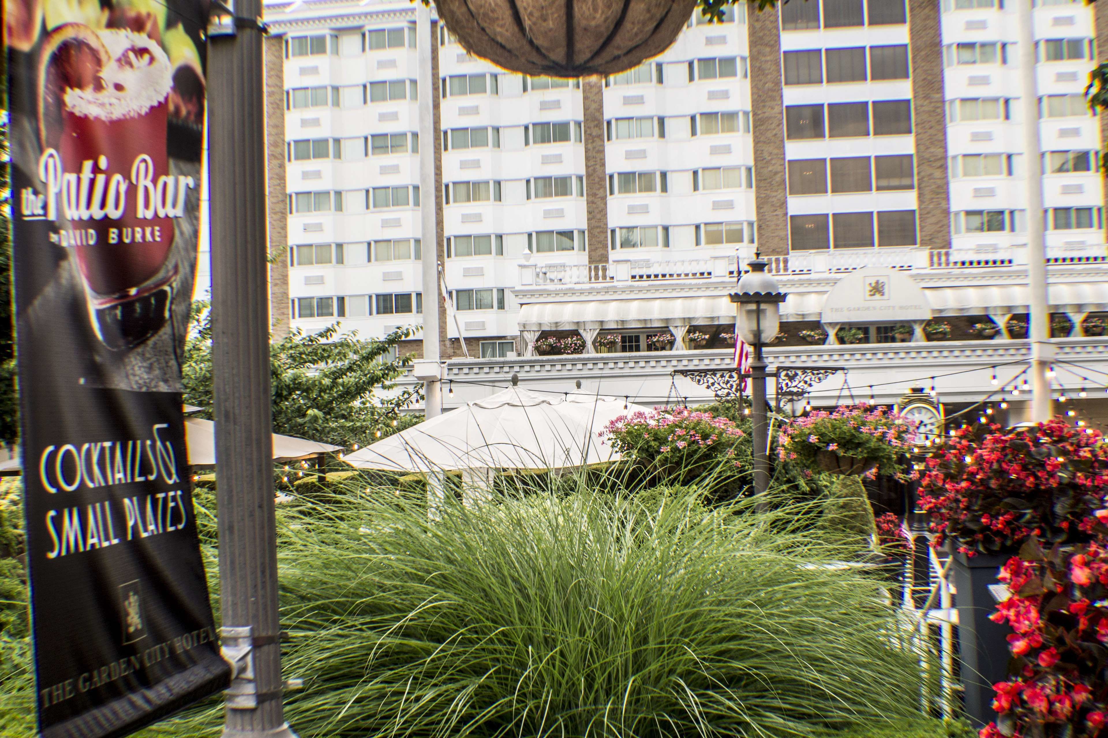 Un hôtel avec une grande façade, un jardin avec de l’herbe haute et des fleurs, et une enseigne indiquant « The Patio Bar ».