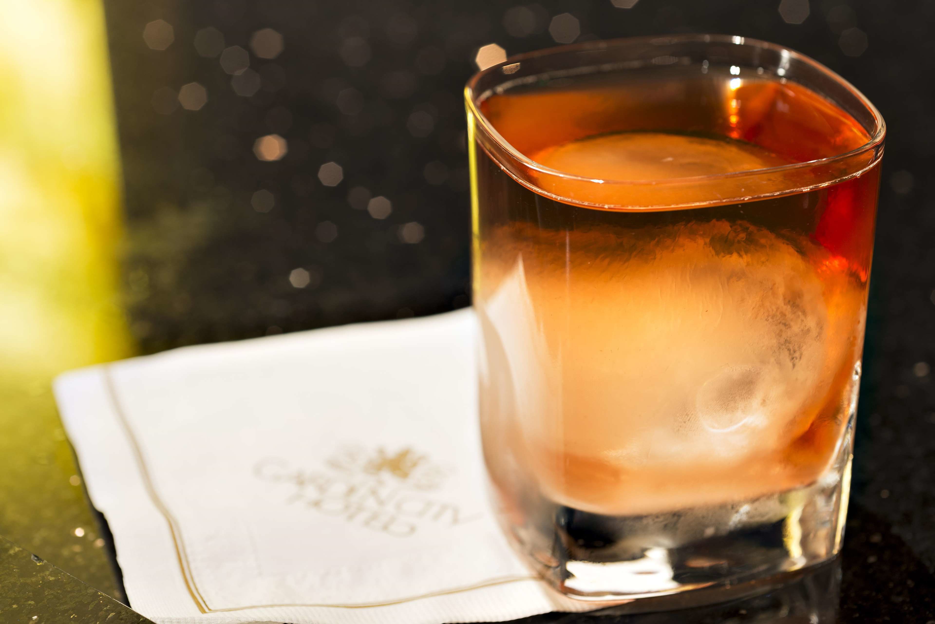 A glass of amber-colored liquid with ice cubes, placed on a white napkin with a logo, on a reflective surface.