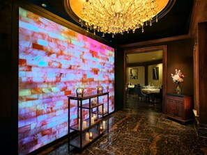 A lobby with a digital wall display, a chandelier, a reception desk, and a dining area in the background. - The Garden City Hotel (Garden City)
