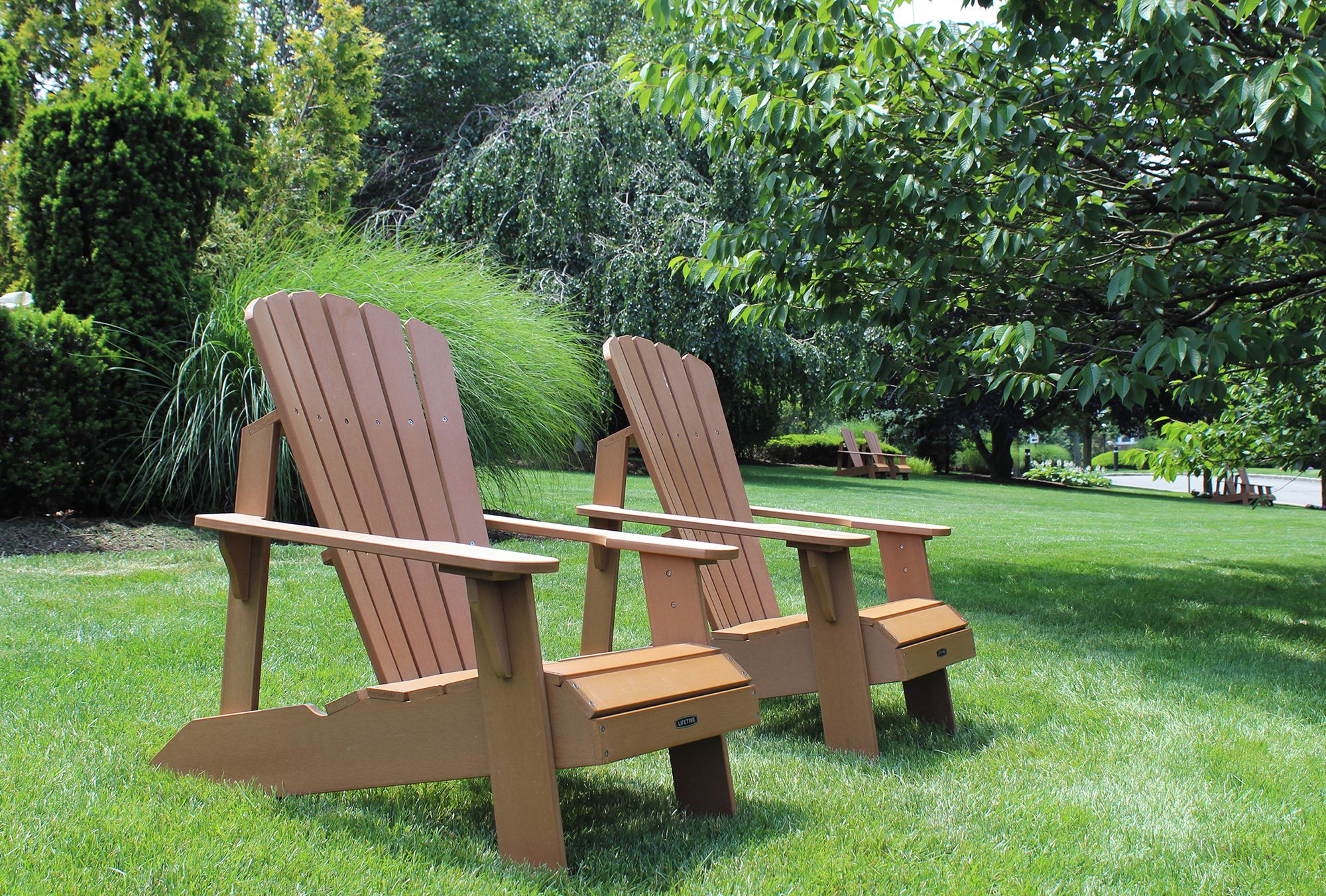 Dos sillas Adirondack de madera con reposabrazos y un banco, ubicados en un césped bien cuidado con arbustos y árboles variados al fondo.
