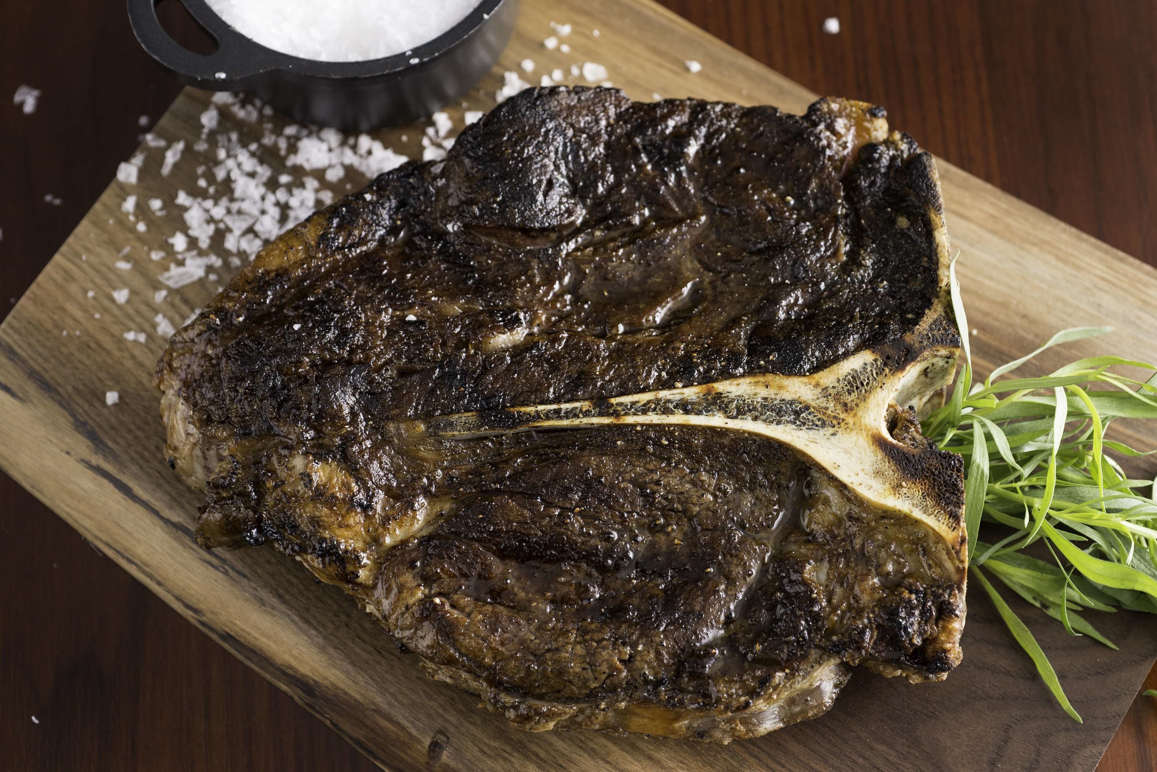 A grilled steak on a wooden board with a sprig of rosemary and a bowl of salt.
