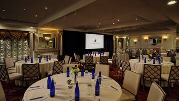Una sala conferenze di un hotel con tavoli rotondi, sedie e uno schermo di proiezione che mostra il nome dell'hotel.