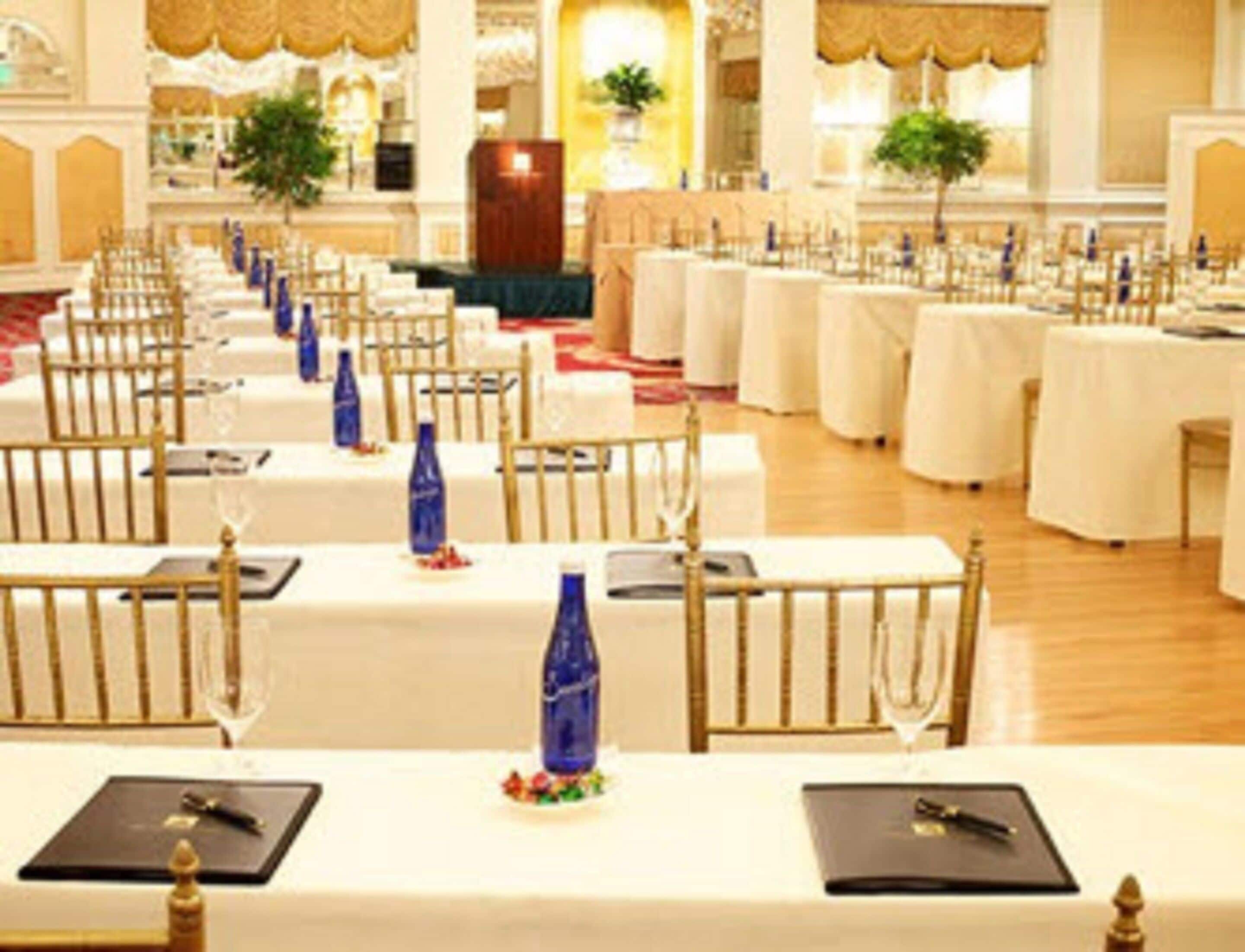 A banquet hall with rows of tables set for a formal event, featuring white tablecloths, gold chairs, and blue water bottles.