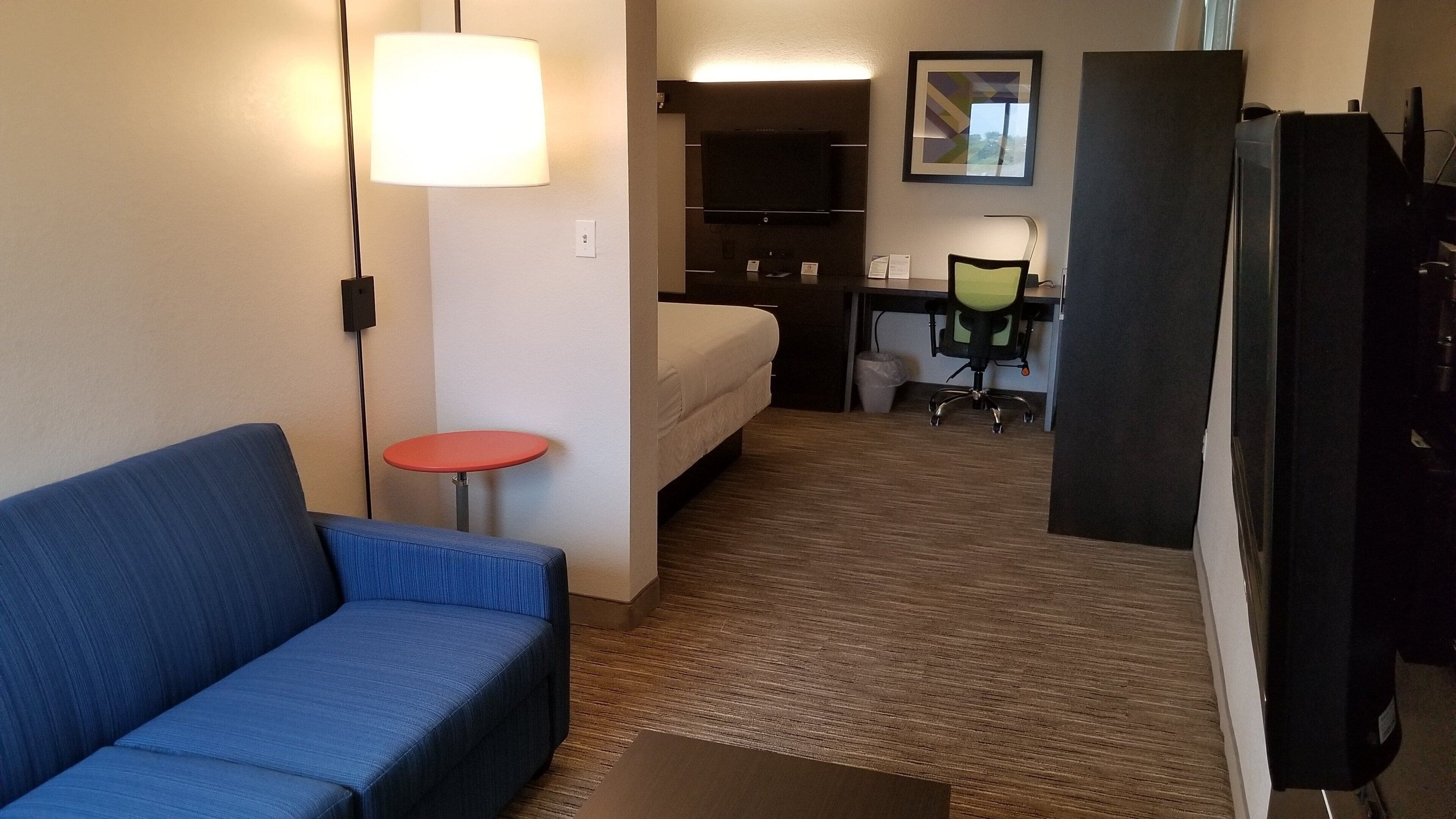 Et hotelværelse med en blå sofa, et lille rødt bord, en seng, et skrivebord med stol, et fjernsyn og et køleskab.