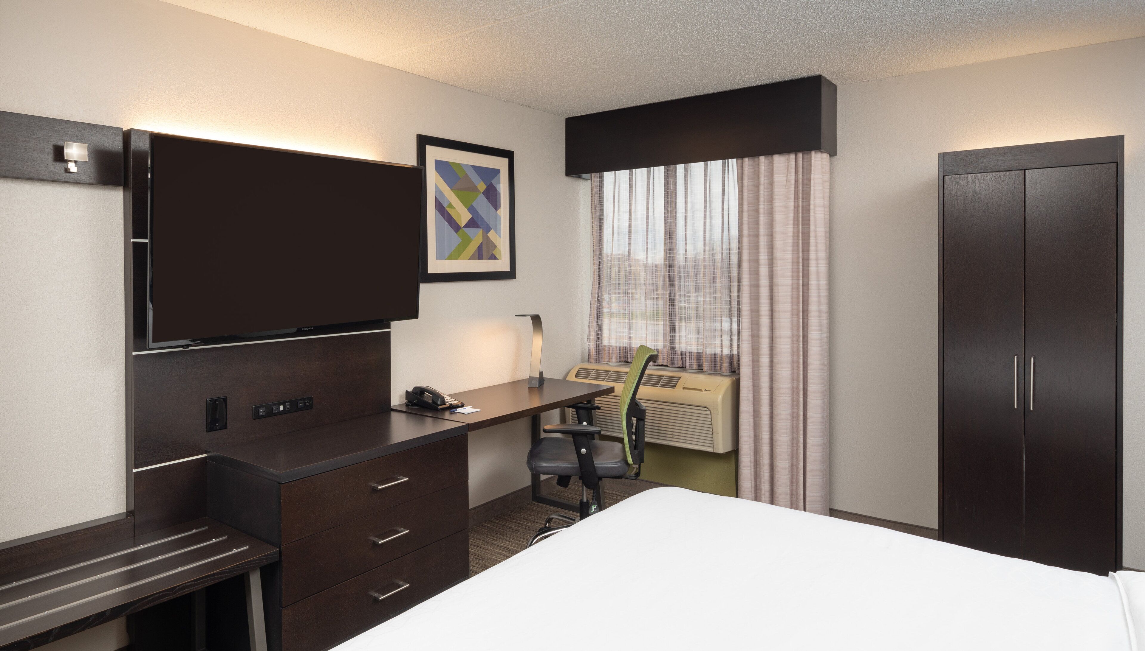 Une chambre d’hôtel comprenant un lit, un bureau avec des tiroirs, une chaise, une télévision, un tableau encadré, une fenêtre avec des rideaux et une armoire.