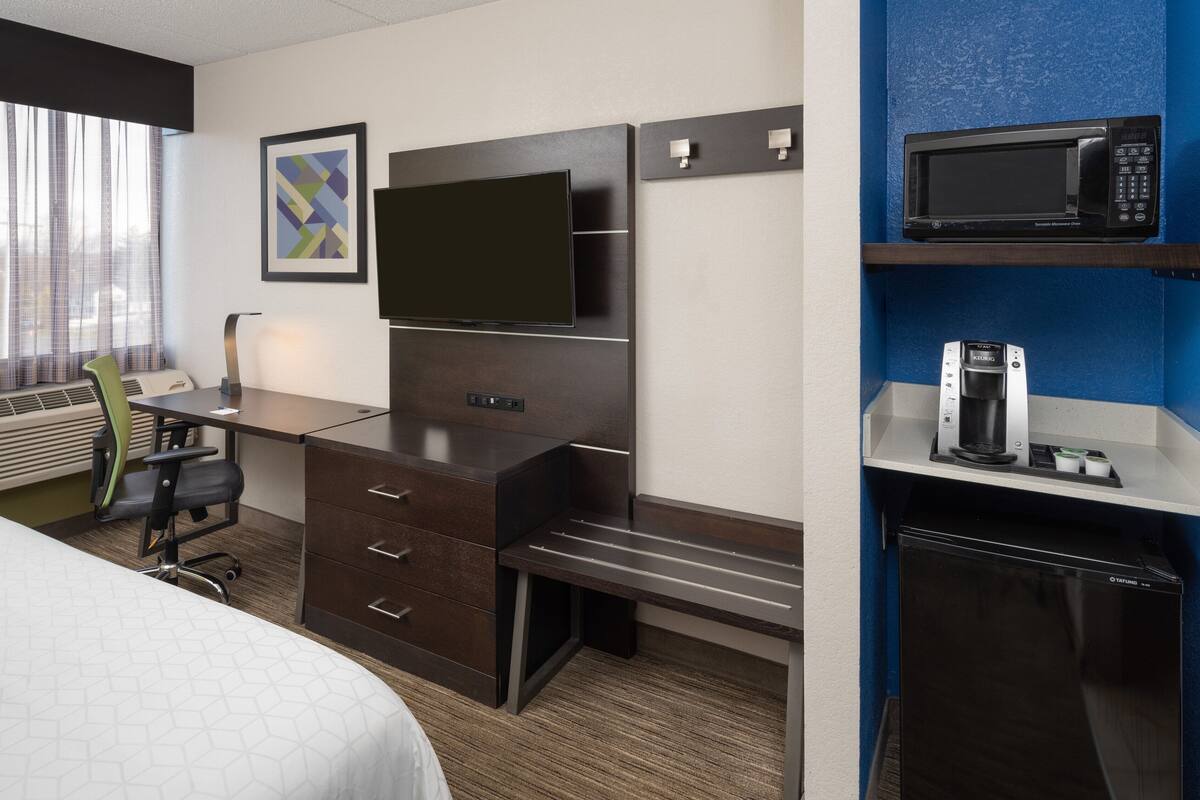 A hotel room with a bed, a desk with a chair, a flat-screen TV, a minibar, and a window with curtains.