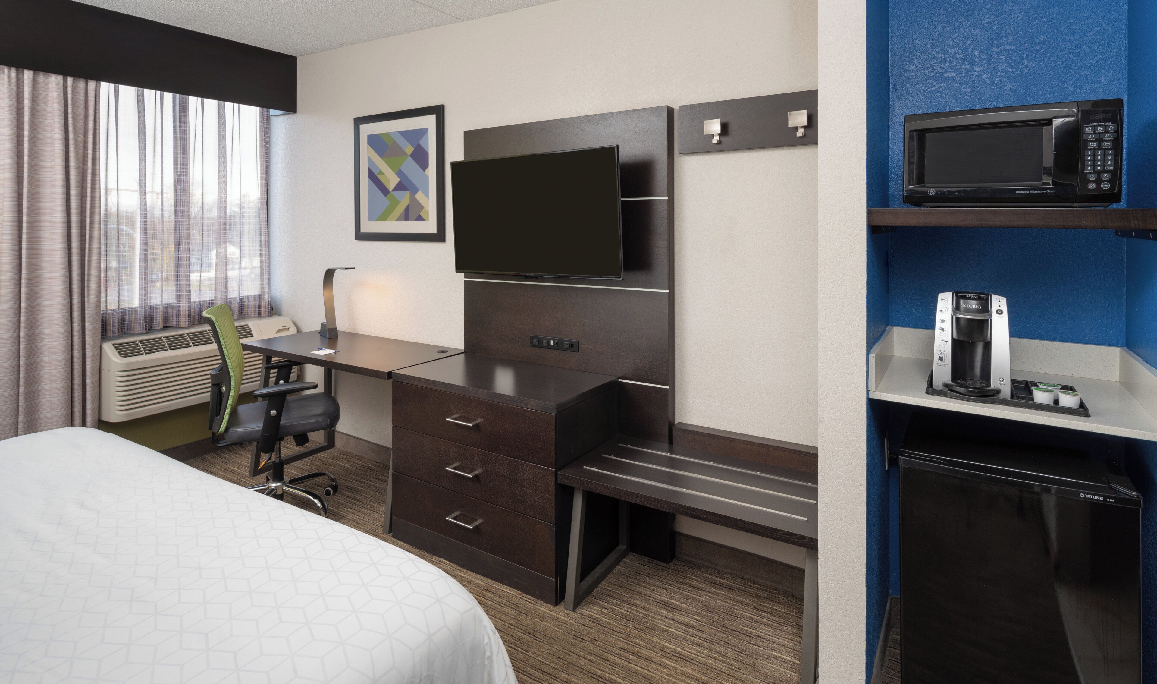 A hotel room with a bed, a desk with a chair, a flat-screen TV, a minibar, and a window with curtains.