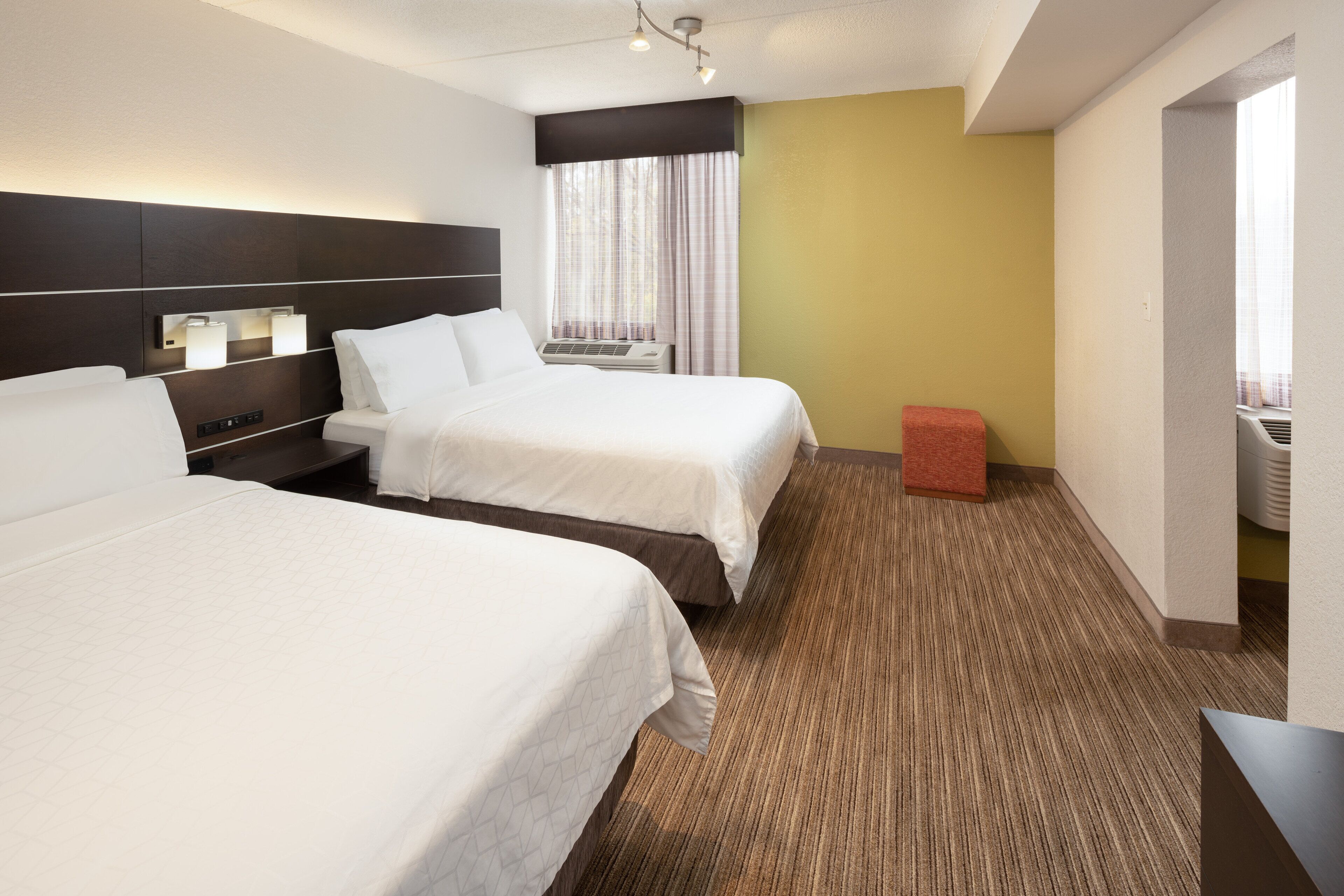 A hotel room with two beds, a small red stool, a window with curtains, and a wall-mounted air conditioning unit.