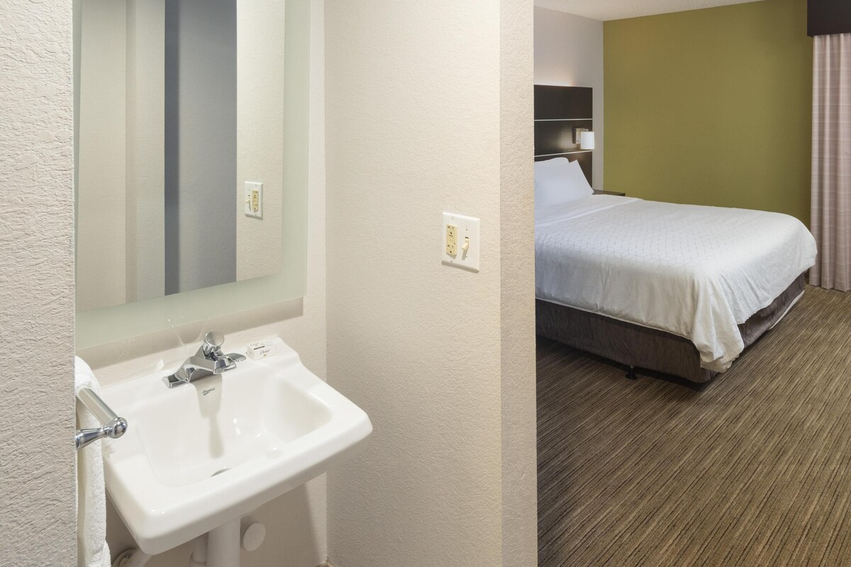 A hotel room with a white sink, a mirror, a bed with a white bedspread, and a green wall.