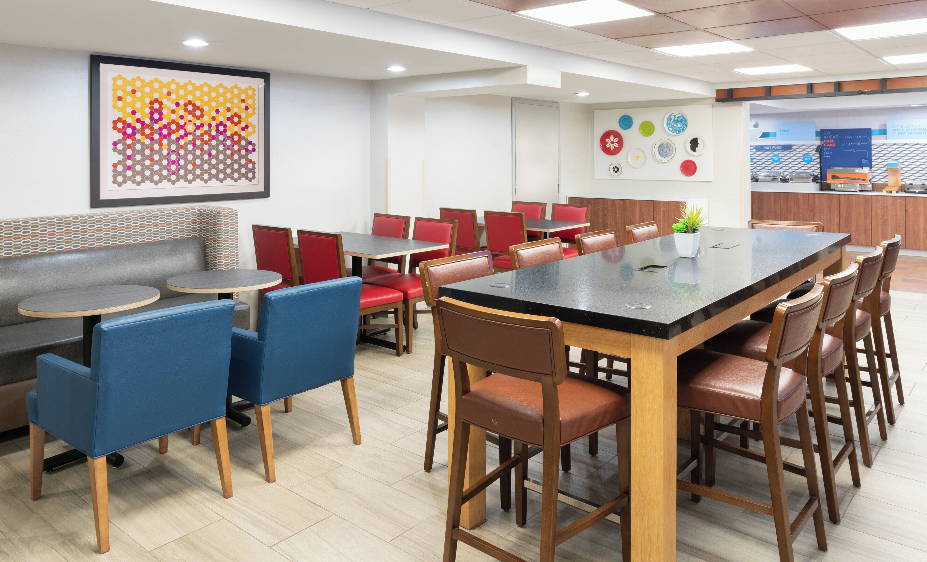 A modern restaurant interior with round tables, red and blue chairs, a long communal table, and a colorful abstract artwork on the wall.