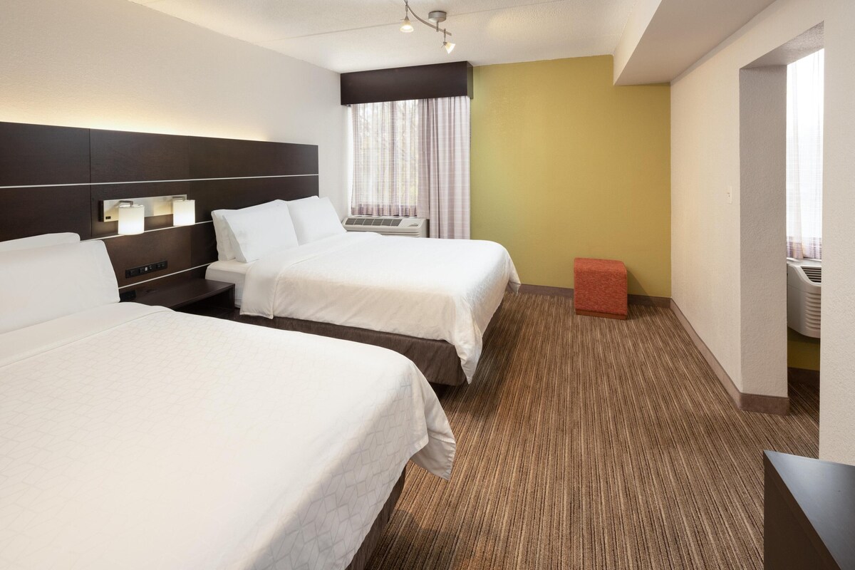 A hotel room with two beds, a small red ottoman, a headboard with built-in lighting, and a window with curtains.