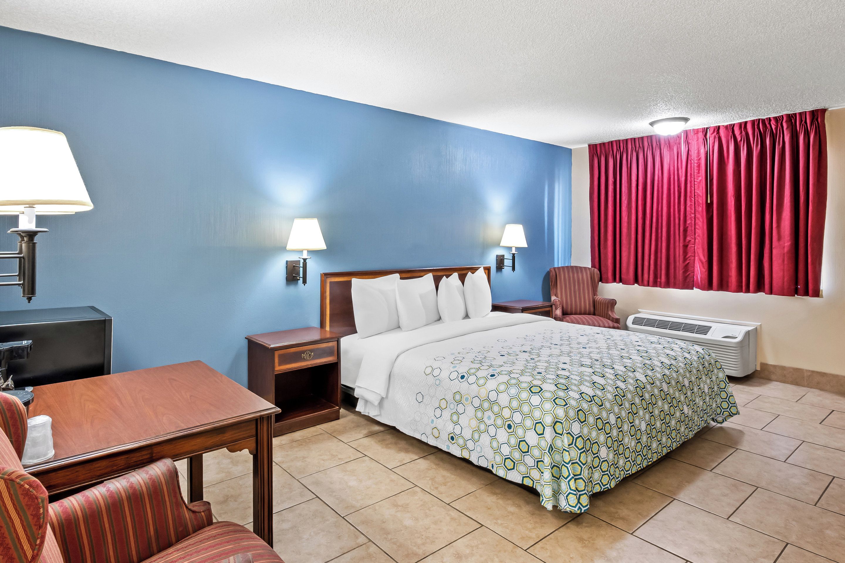 A hotel room with a bed, a desk, a chair, two bedside lamps, a window with red curtains, and an air conditioning unit.