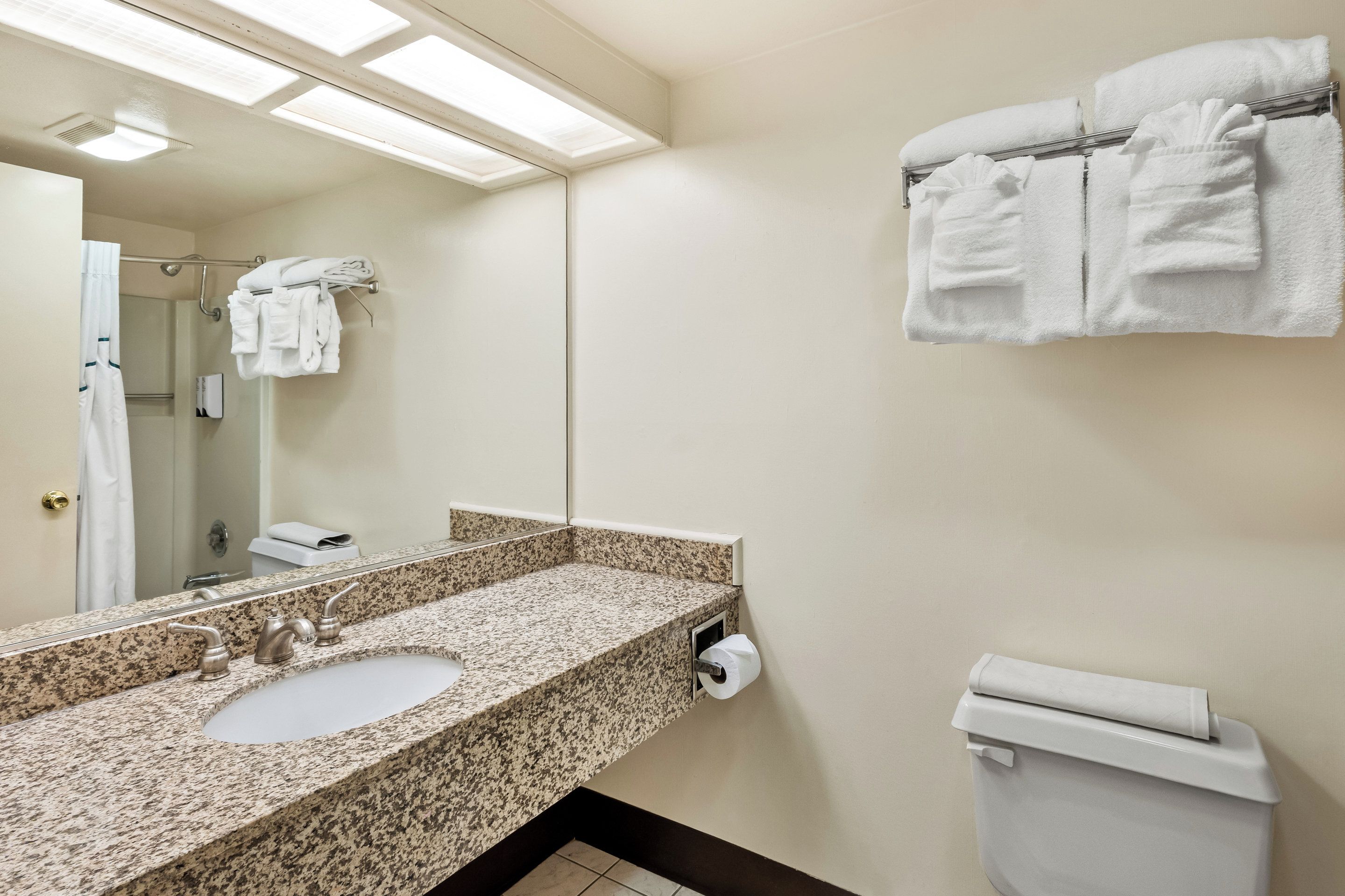 A bathroom with a granite countertop sink, a mirror, a towel rack with folded towels, a toilet, and a paper towel dispenser.