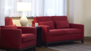 Een kamer met een rode bank, een kleinere fauteuil, een bijzettafeltje met een lamp en een raam met dunne gordijnen.