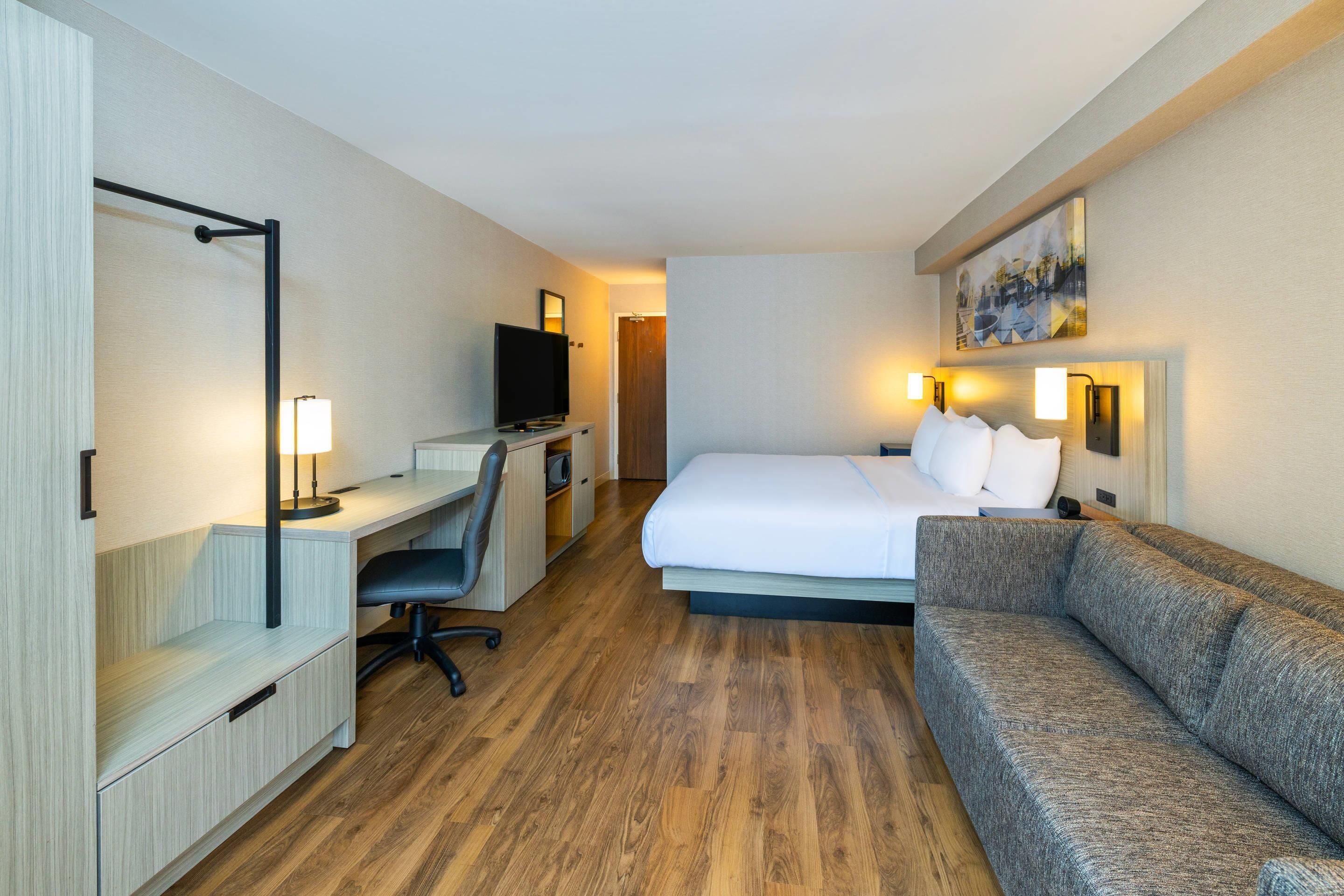 A hotel room with a bed, a sofa, a desk with a computer, a TV, and a painting on the wall.