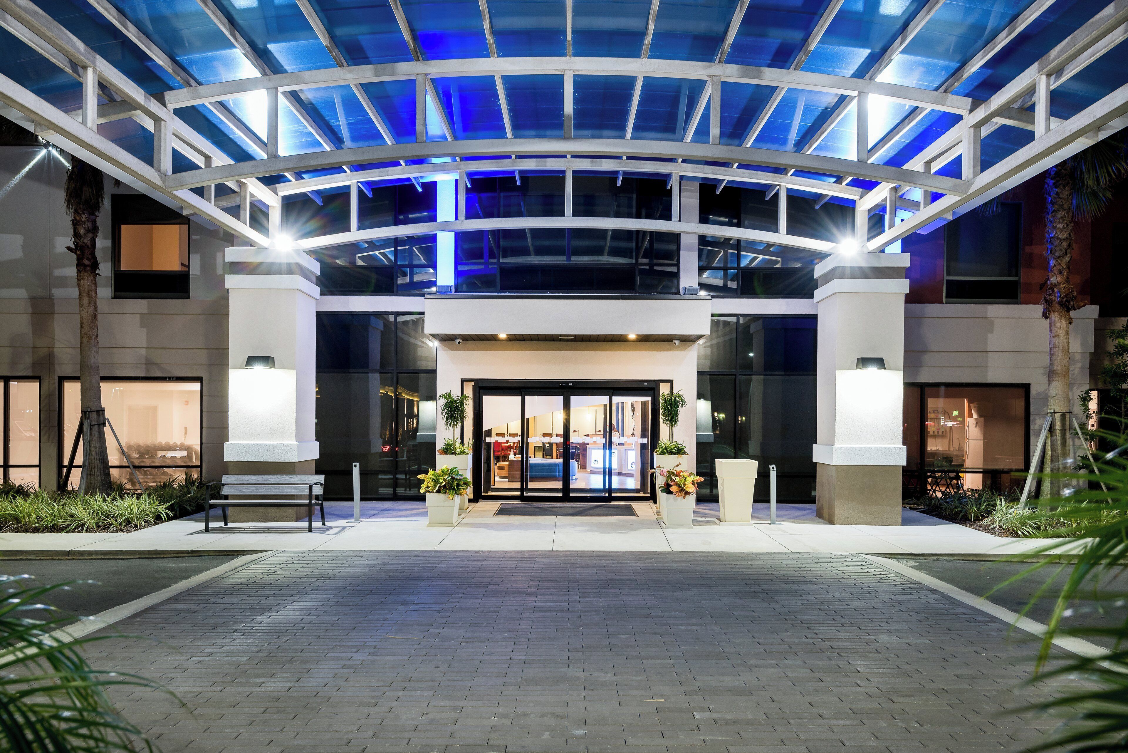 A hotel entrance with a glass canopy, a reception desk, and a seating area with plants.