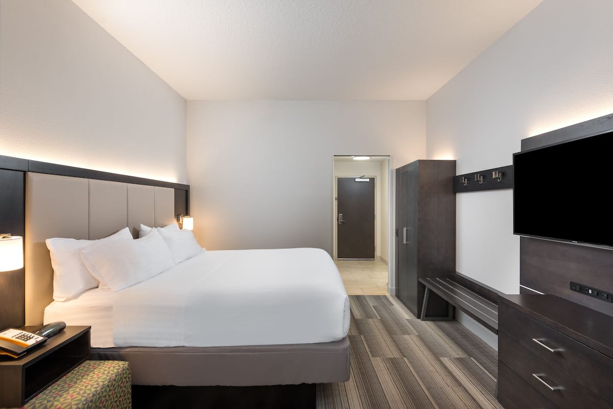 Een hotelkamer met een bed, nachtkastje, lamp, televisie, kledingkast en een vloerbedekking.