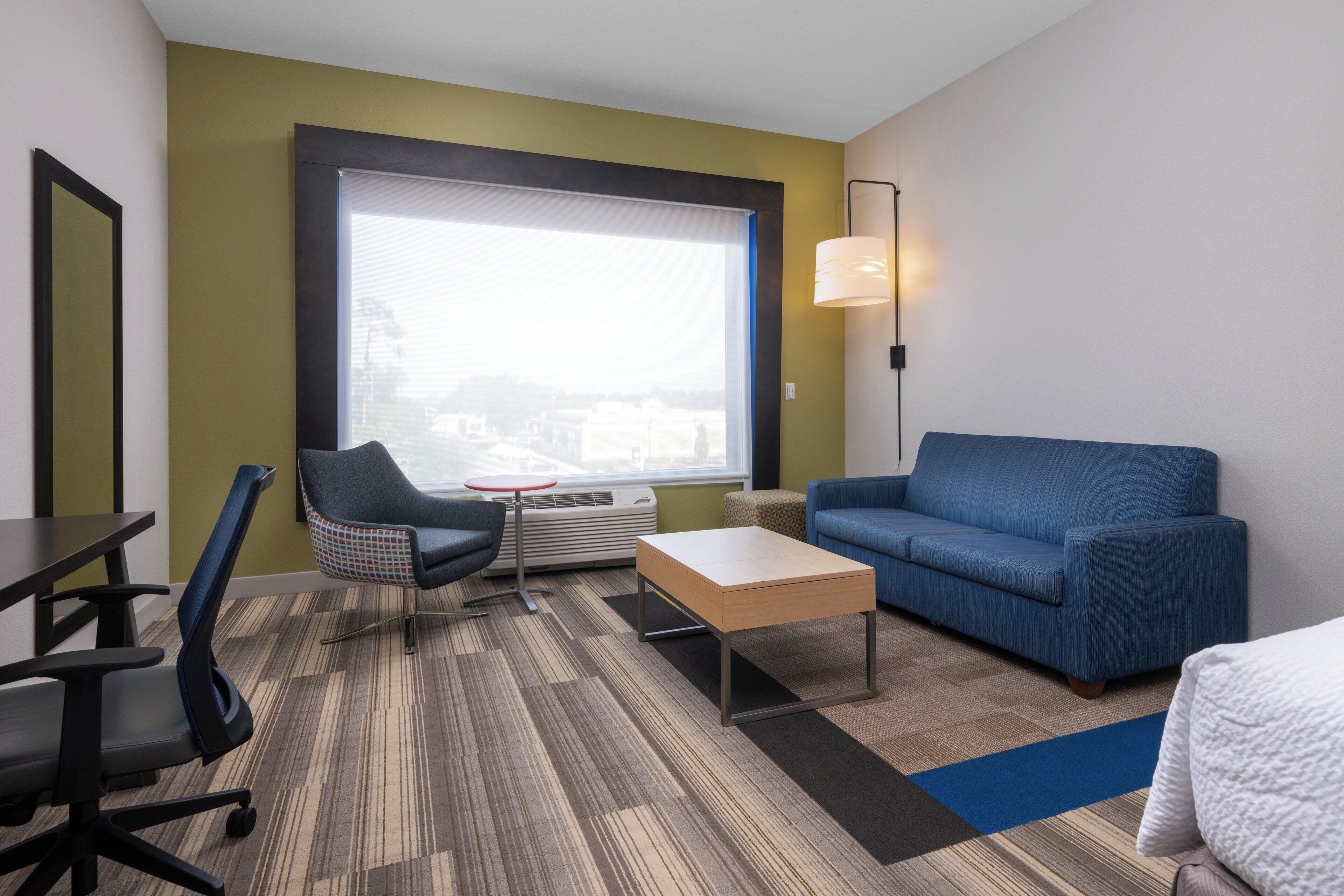 Habitación de hotel con un ventanal grande, un sofá azul, una mesita, una silla y un escritorio.