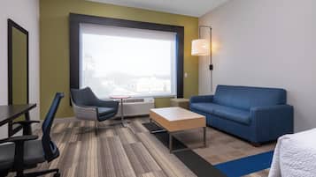 Habitación de hotel con un ventanal grande, un sofá azul, una mesita, una silla y un escritorio.