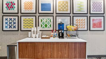 A hotel reception area with a wooden counter, framed artwork on the wall, and various amenities like coffee, snacks, and toiletries.