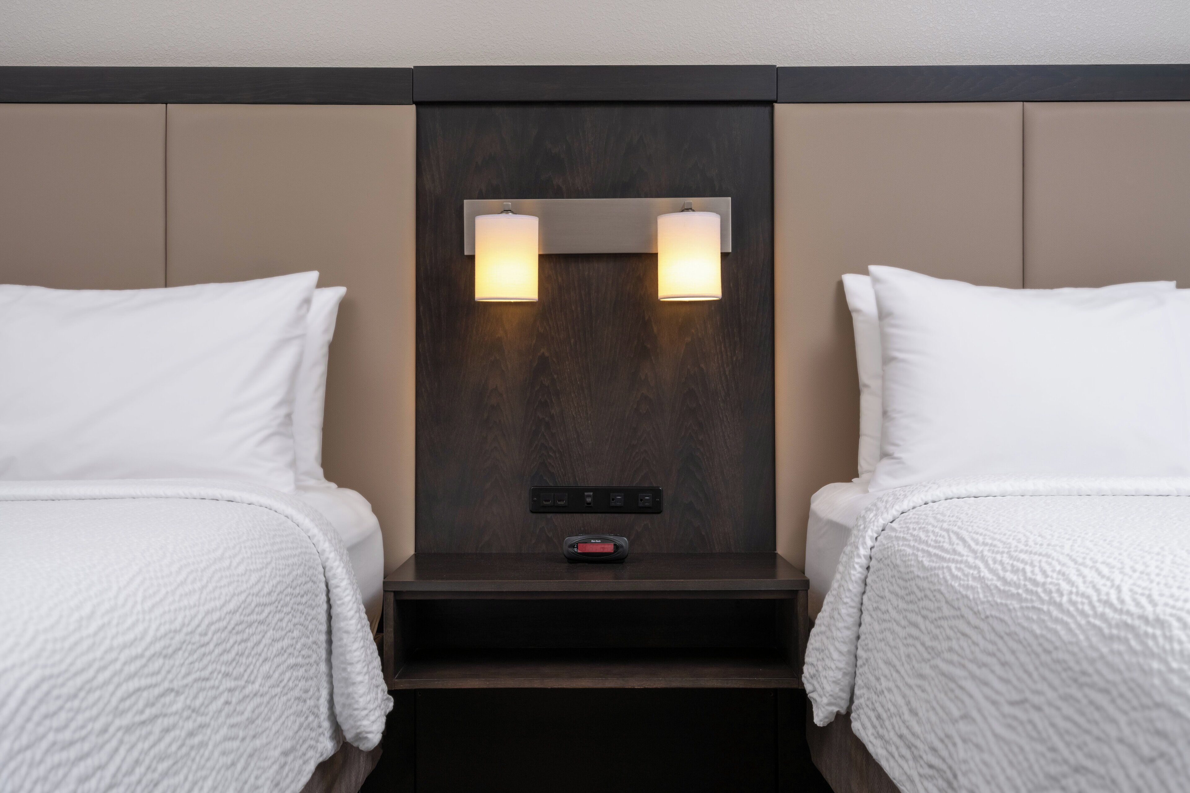 Habitación de hotel con dos camas, una mesita de noche con panel de control e iluminación en la pared.