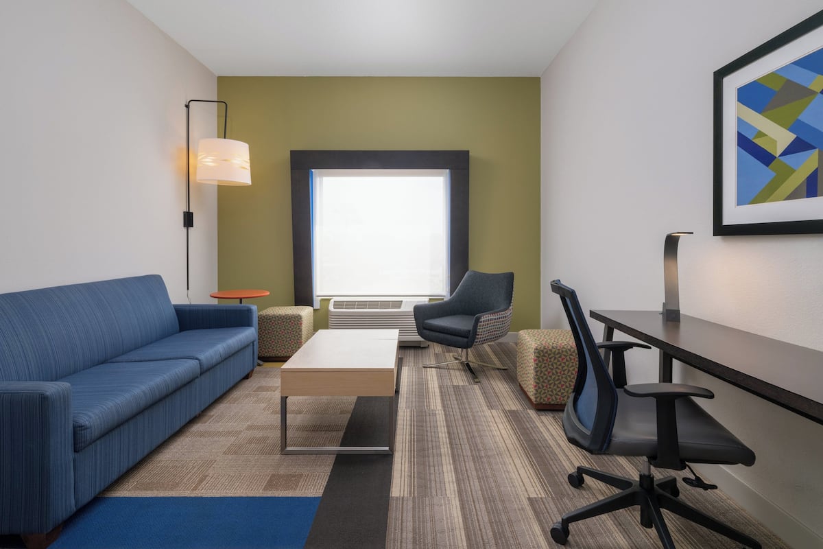 Een moderne hotelkamer met een blauwe bank, een kleine ronde tafel, een groot raam, een stoel en een bureau.
