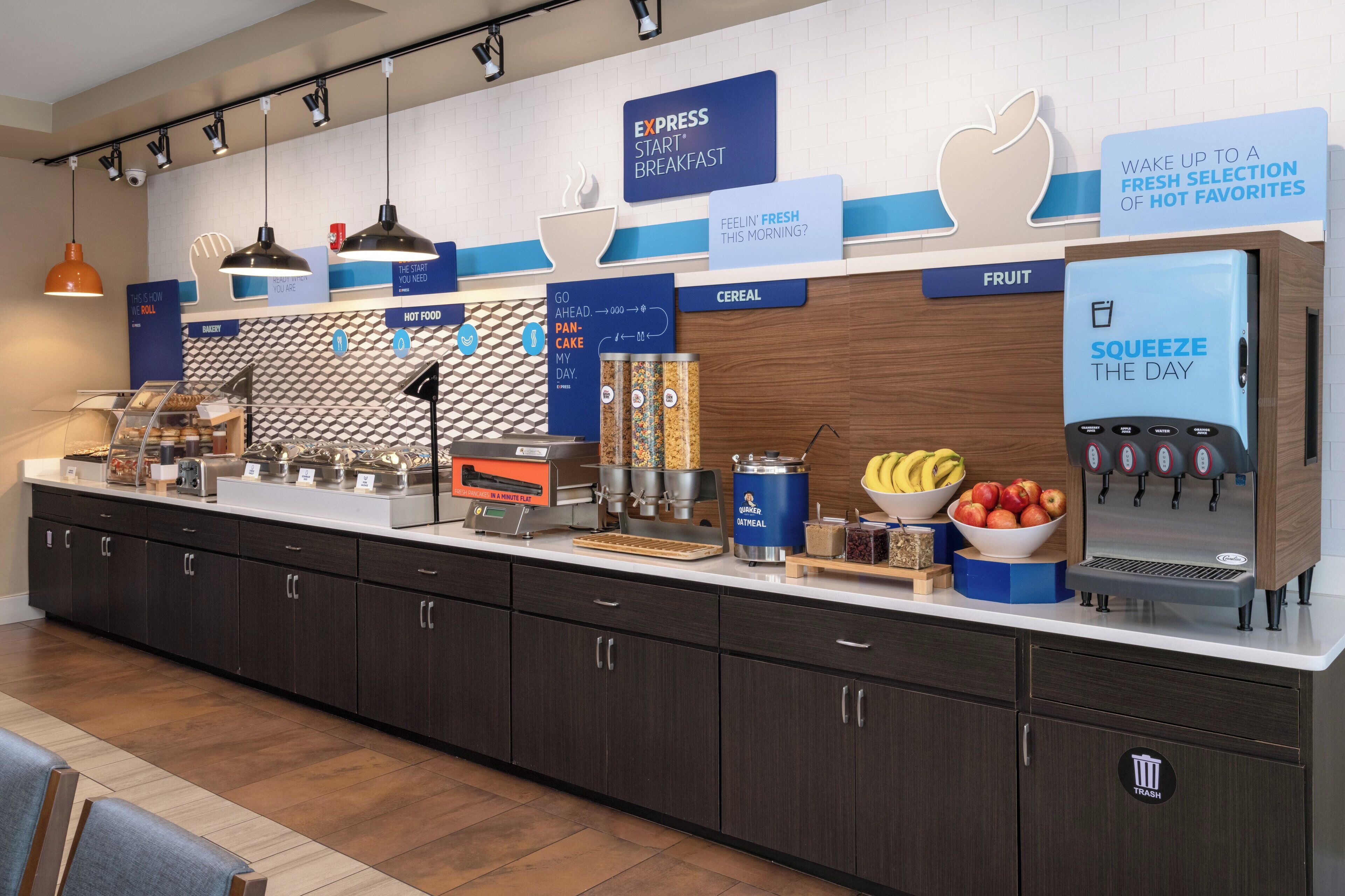 Una barra de desayuno con diversas opciones de comida, como cereales, fruta y alimentos calientes. Se observan carteles que indican la disponibilidad de desayuno exprés y un dispensador de zumo.