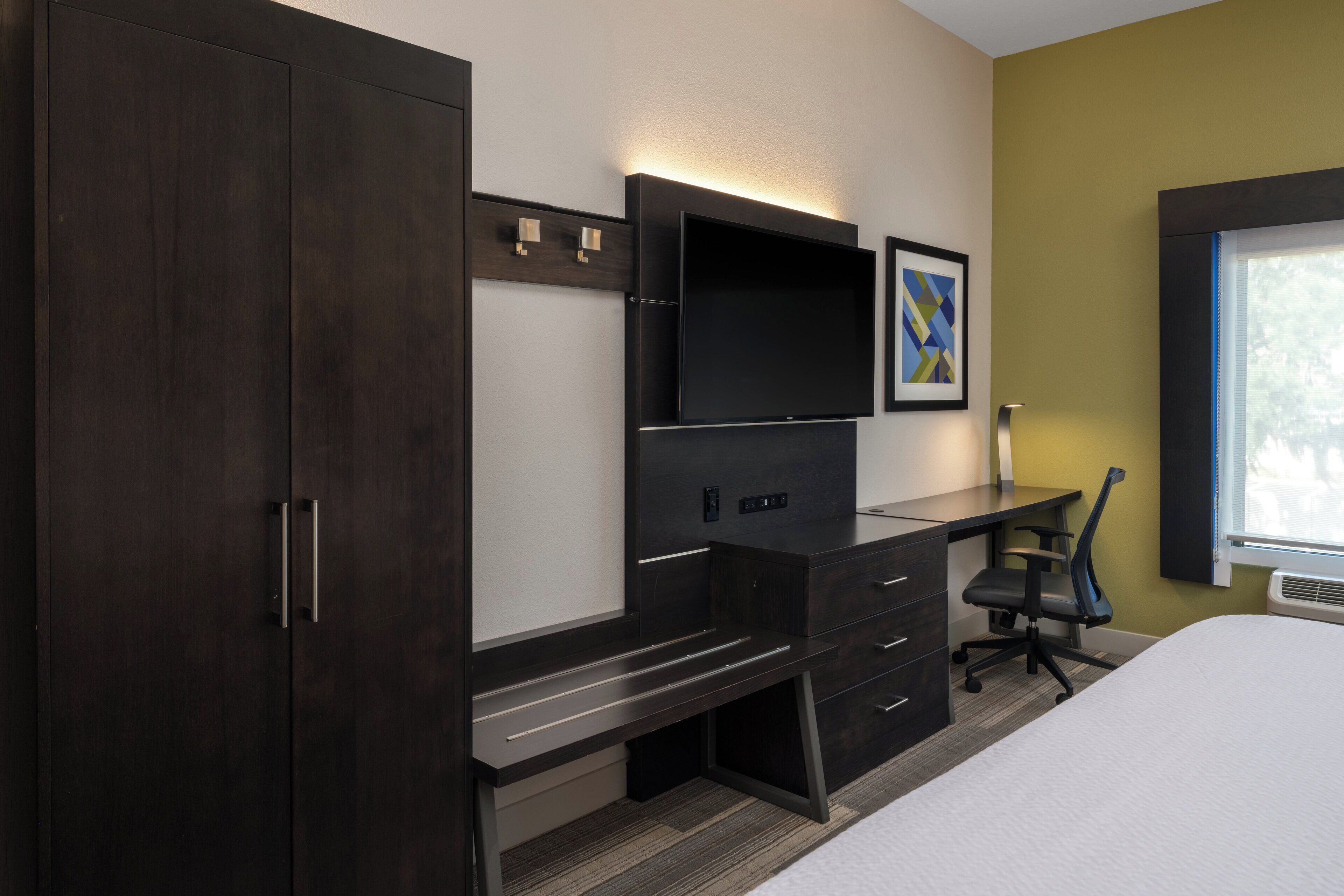 Een slaapkamer met een bed, een bureau met stoel, een televisie en een kledingkast.