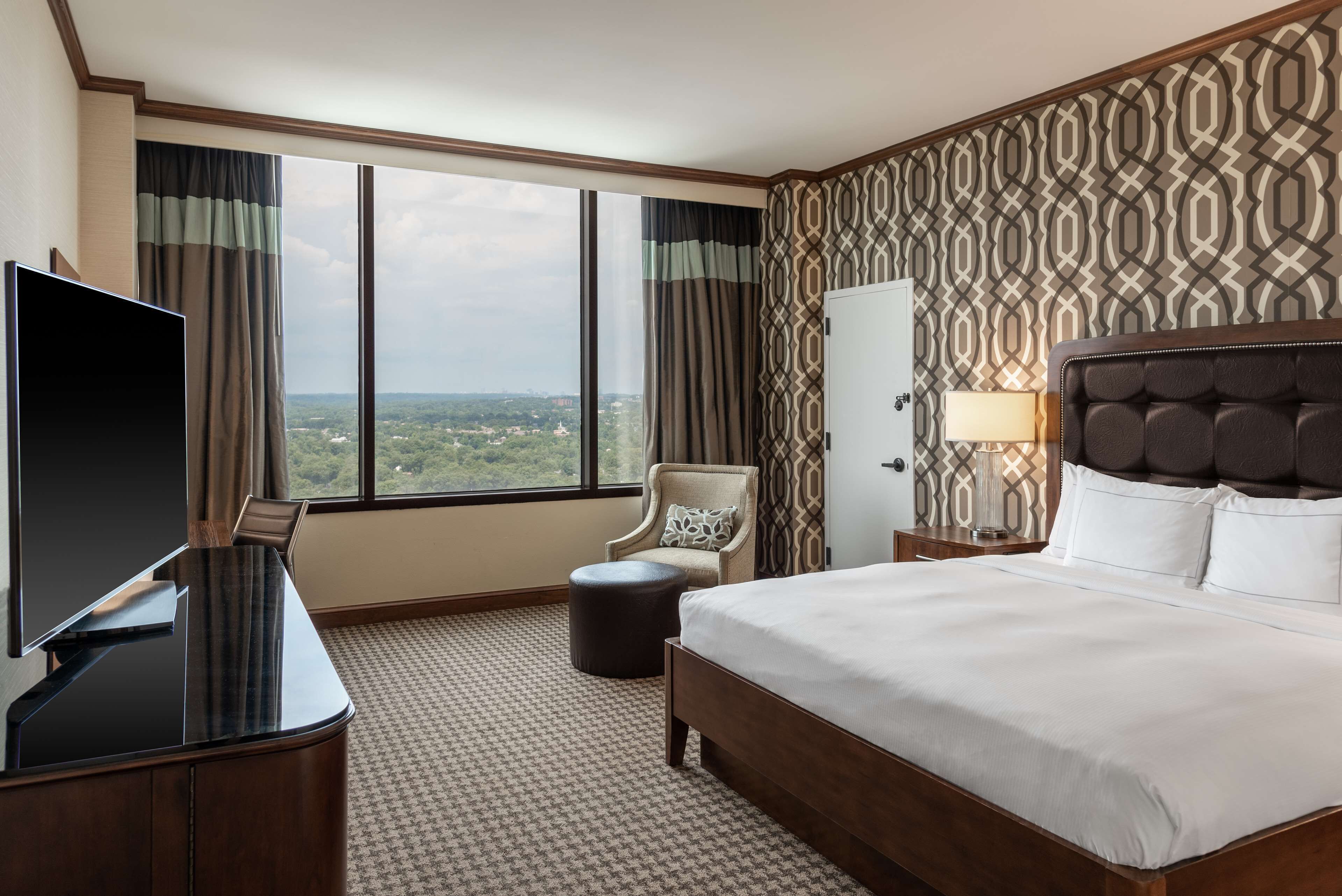 Habitación de hotel con una cama grande, televisión, sillón y vista a la ciudad a través de ventanas amplias.