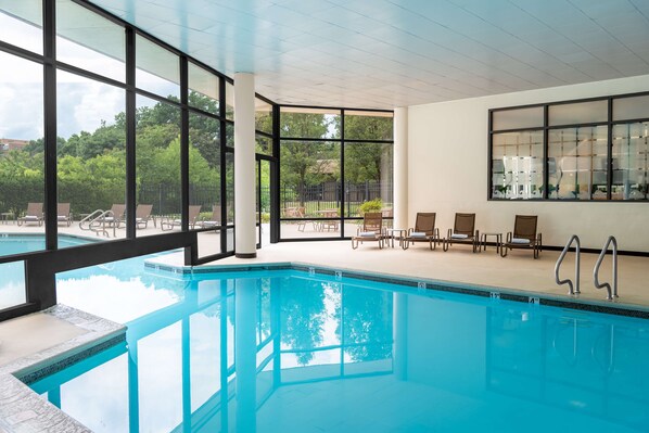 A swimming pool with clear blue water, surrounded by lounge chairs and a view of greenery outside.