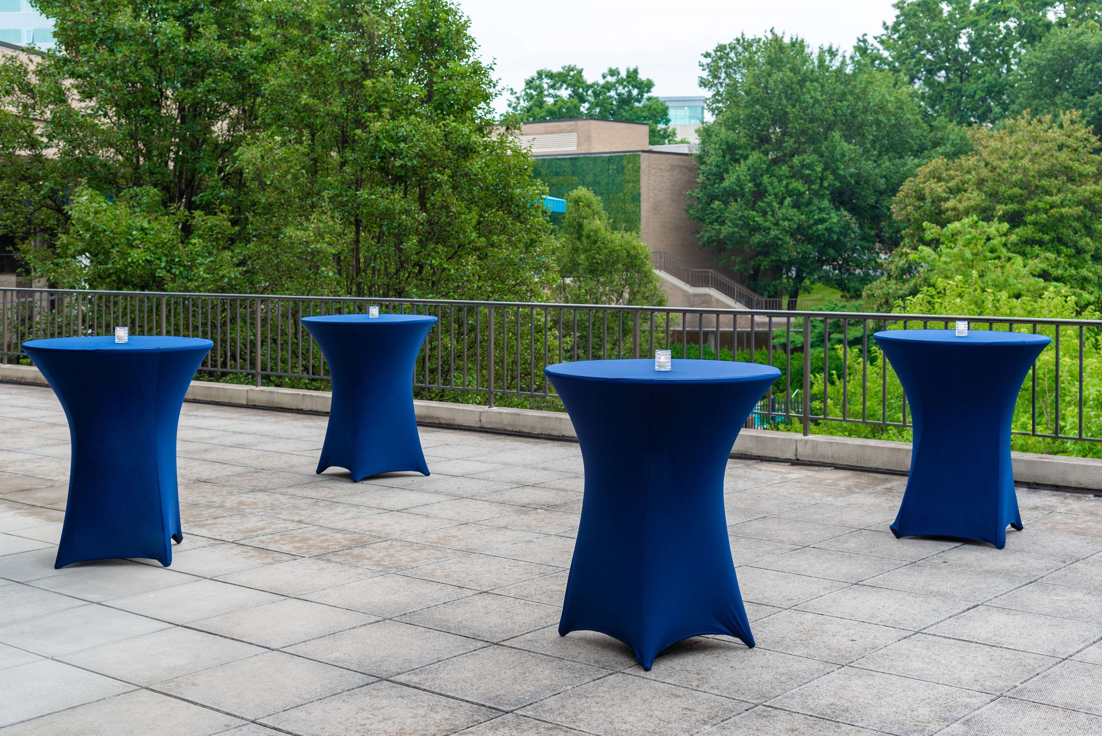 Drei blaue Tische draußen auf einer gefliesten Terrasse mit Metallgeländer und Bäumen im Hintergrund.