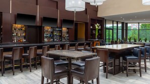 A modern bar area with a long counter, multiple seating options, and a large window providing a view of greenery. - Hilton Alexandria Mark Center (Alexandria)