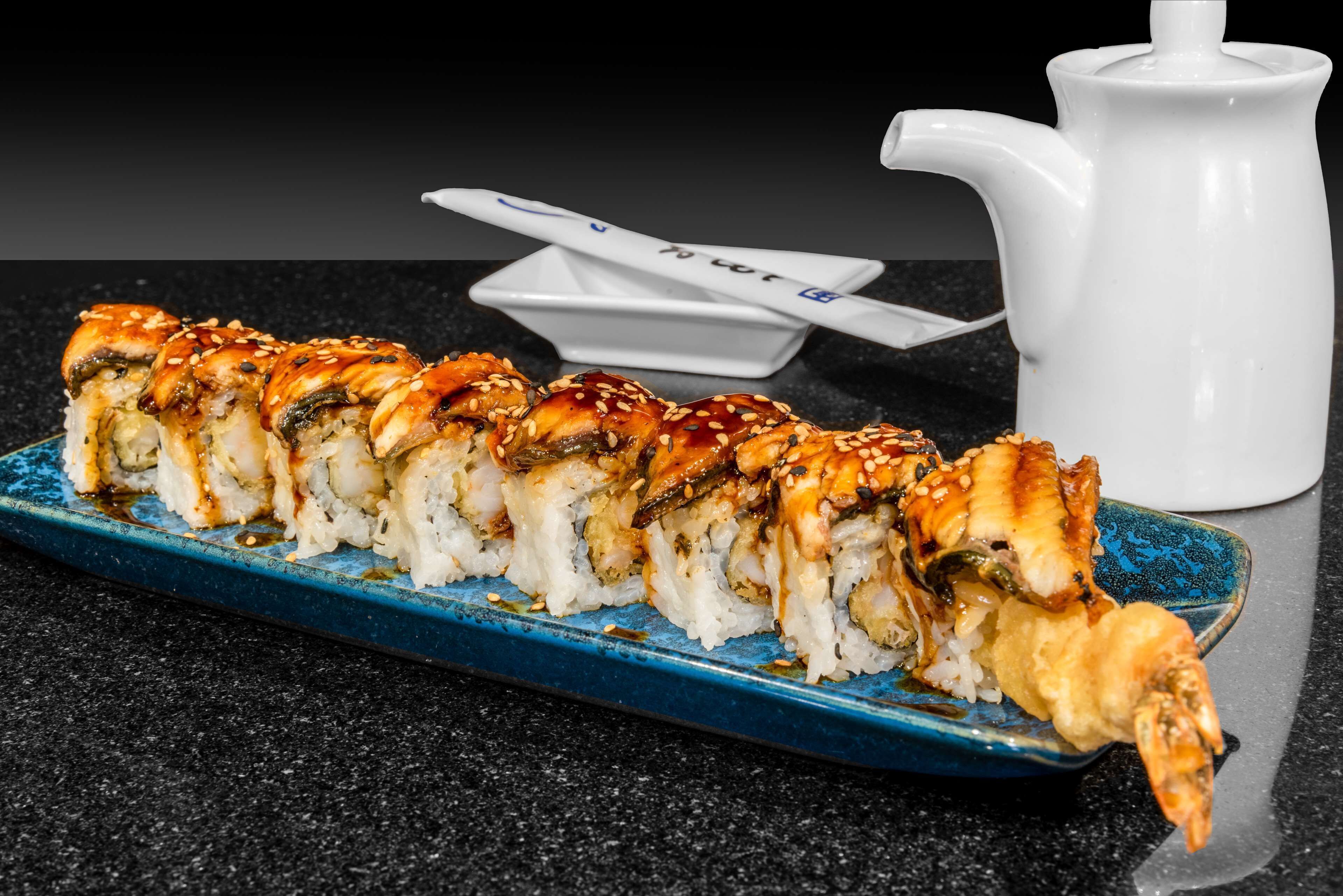 Een sushi bord op een blauw bord, met een witte karaf en stokjes op de achtergrond.