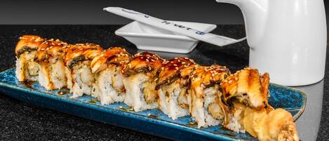 Een sushi bord op een blauw bord, met een witte karaf en stokjes op de achtergrond.
