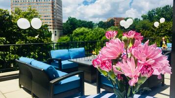 Un espace extérieur aménagé avec des meubles en osier et des coussins bleus, une table recouverte d’une nappe à rayures et un vase rempli de fleurs roses.