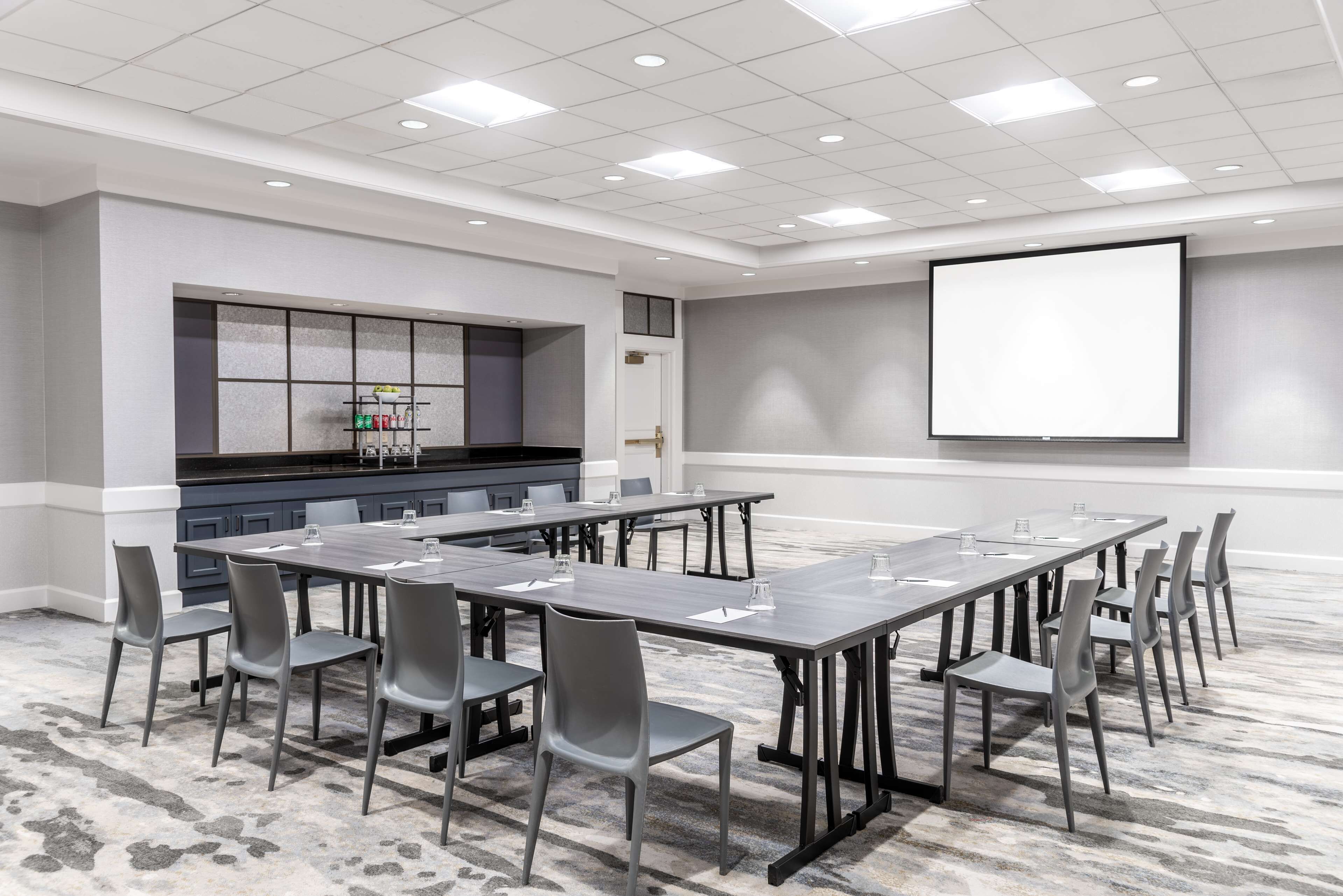 A conference room with a long table, chairs, a projector screen, and a kitchenette area.